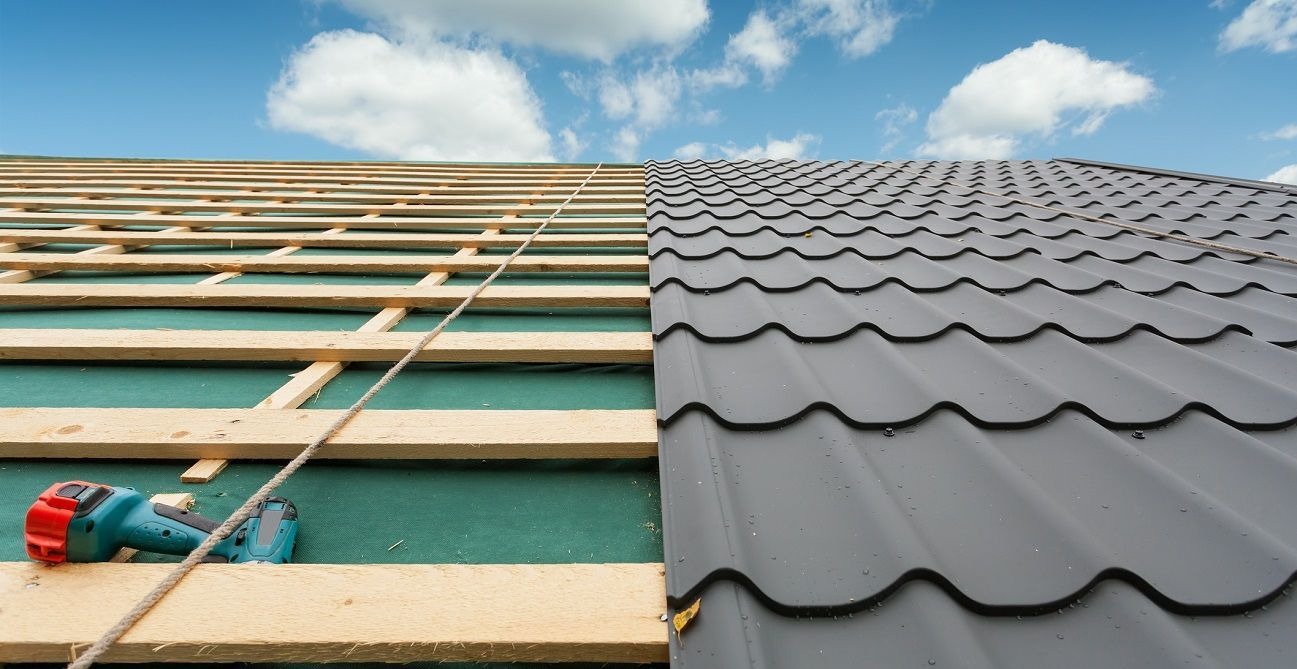 Połowa dachu pokryta szarą blachodachówką, druga połowa z drewnianą konstrukcją i wkrętarką na zielonej membranie, na tle błękitnego nieba.