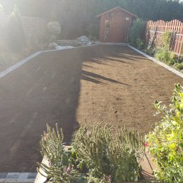 ExtraOgród usługi ogrodnicze - Przygotowany teren pod trawnik z rolki, obrzeża z kostki, w tle drewniany domek narzędziowy i ogrodzenie. Słoneczny dzień.