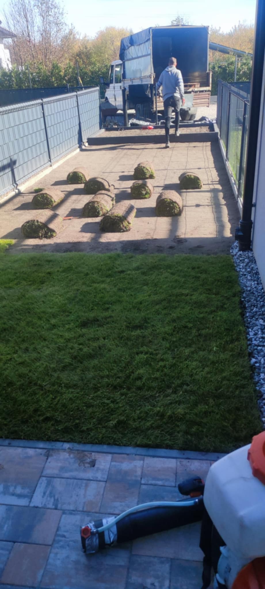 Układanie trawy z rolki w ogrodzie: widoczny fragment ułożonej trawy, rolki trawy na przygotowanym podłożu, pracownik i samochód dostawczy w tle.