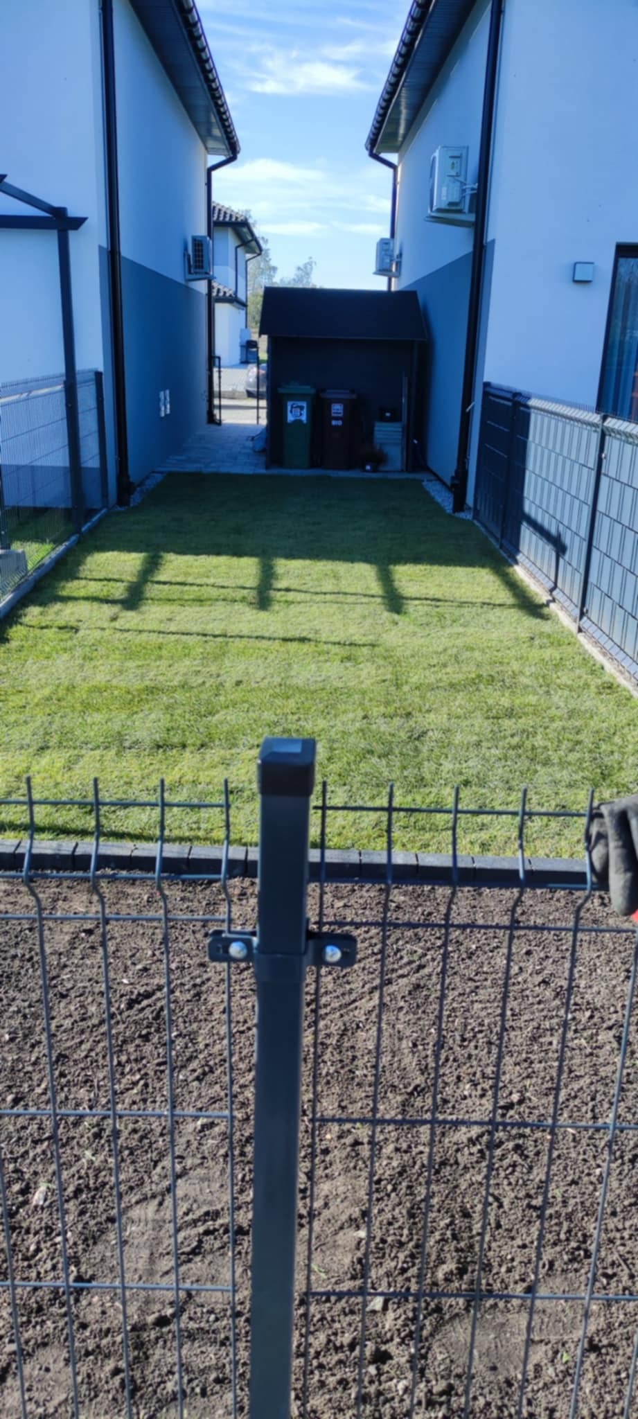 Widok na wąski ogród między domami z nową trawą, widziany przez metalowe ogrodzenie w trakcie montażu. W tle widoczne kosze na śmieci i garaż.