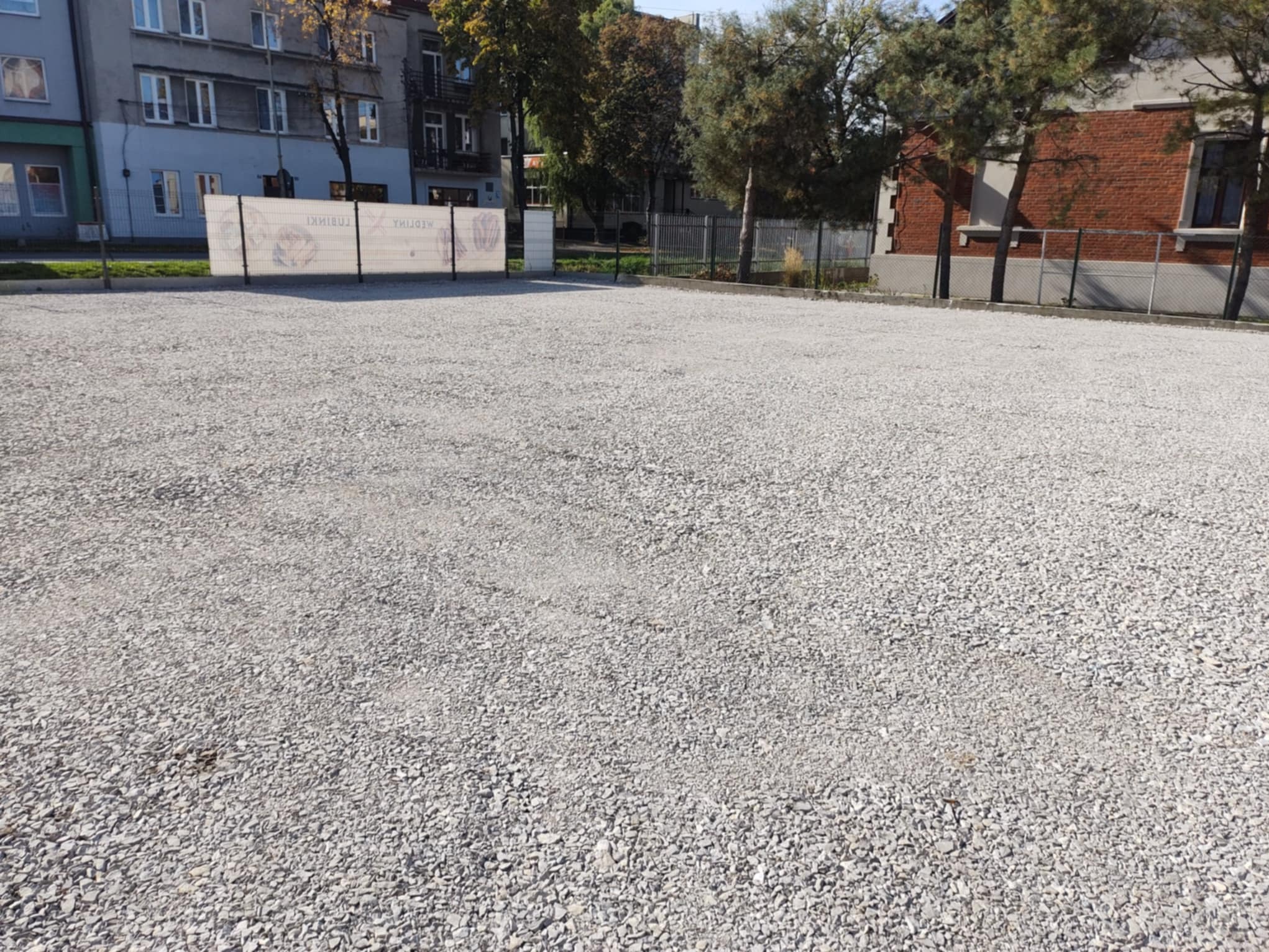 Wyrównany teren pokryty drobnym, szarym żwirem na tle budynków i drzew. Widoczne ogrodzenie z reklamą w tle. Teren przygotowany pod dalsze prace.