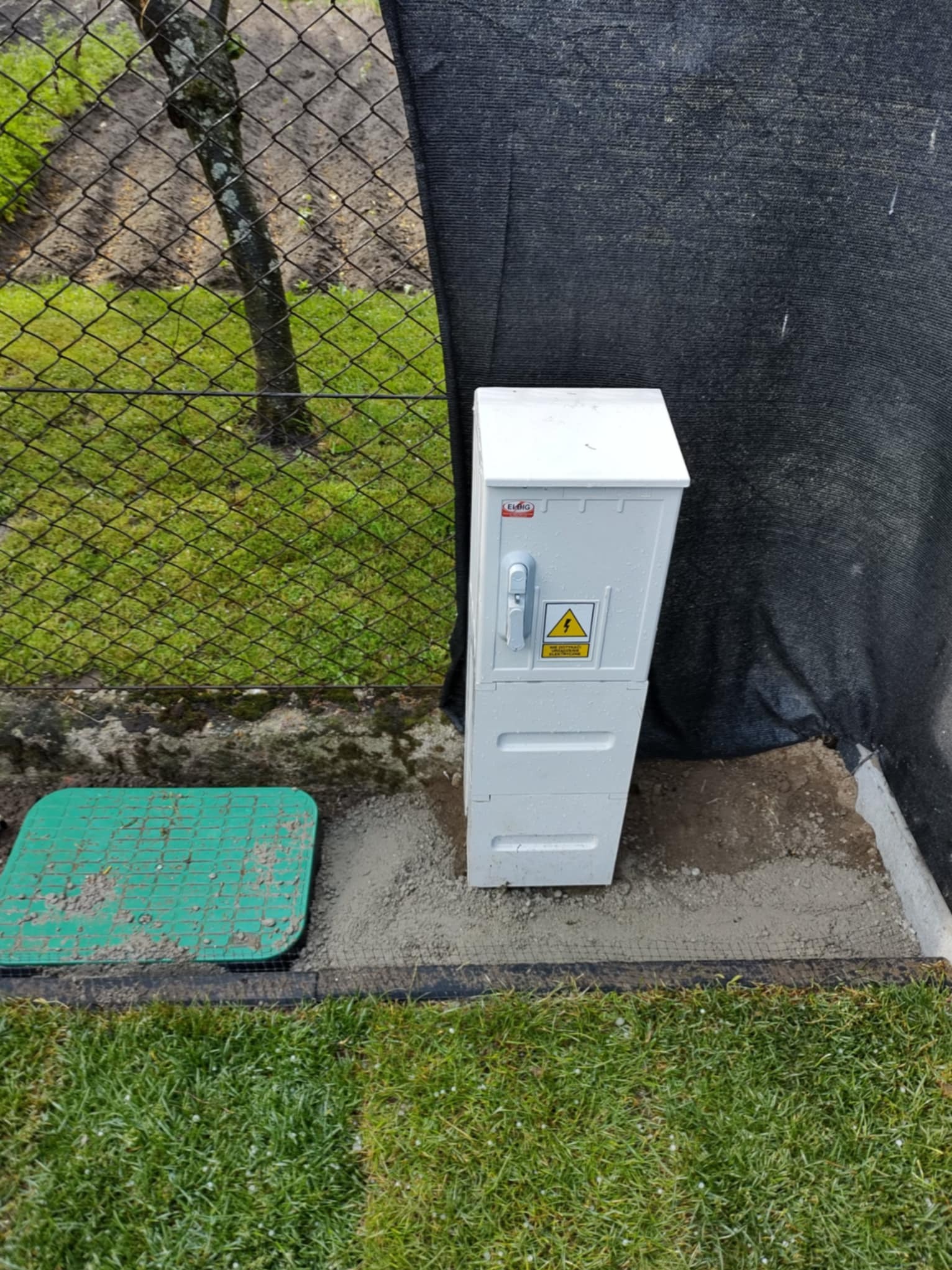 Biała skrzynka elektryczna w ogrodzie w Grabinach, obok zielona pokrywa studzienki, siatka ogrodzeniowa i czarna tkanina. Widoczne świeżo ułożone obrzeże.
