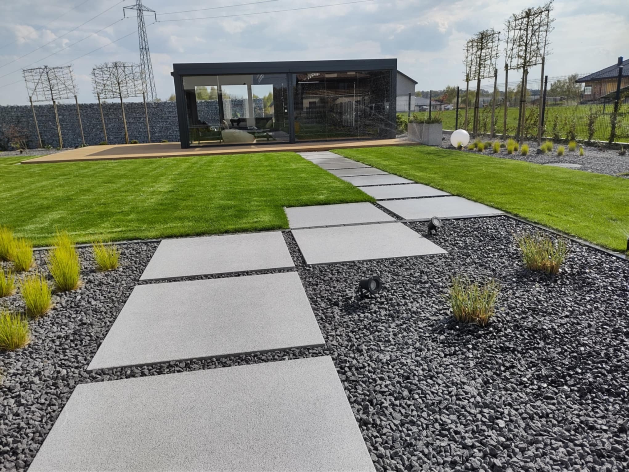 Nowoczesny ogród z równo przystrzyżonym trawnikiem, ścieżką z płyt i szarym żwirem. W tle altana z przeszkleniami i formowane drzewa.