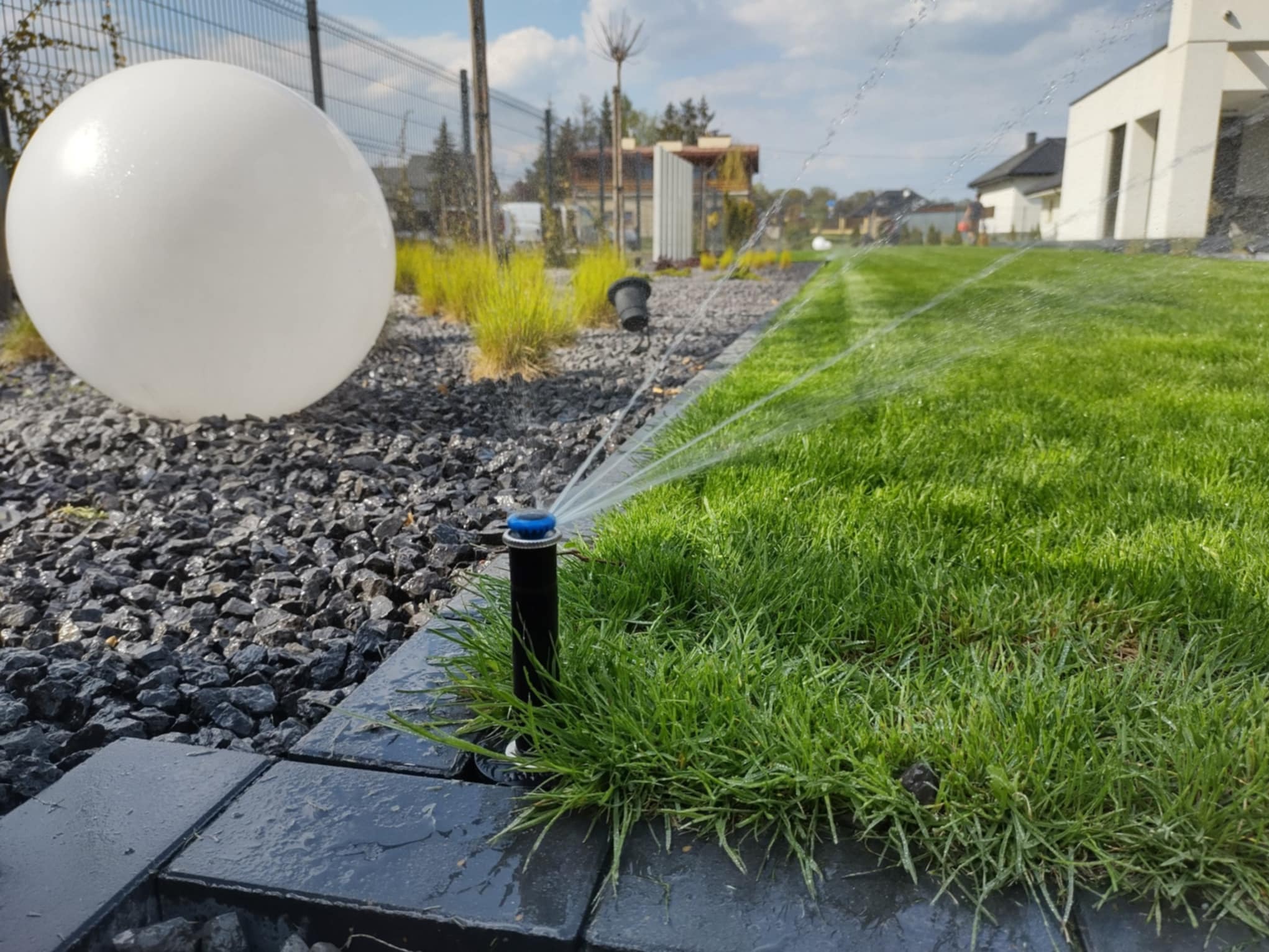 Automatyczny system nawadniania trawnika w ogrodzie z kulistą lampą, zraszacz w akcji, w tle dom jednorodzinny i ogrodzenie.