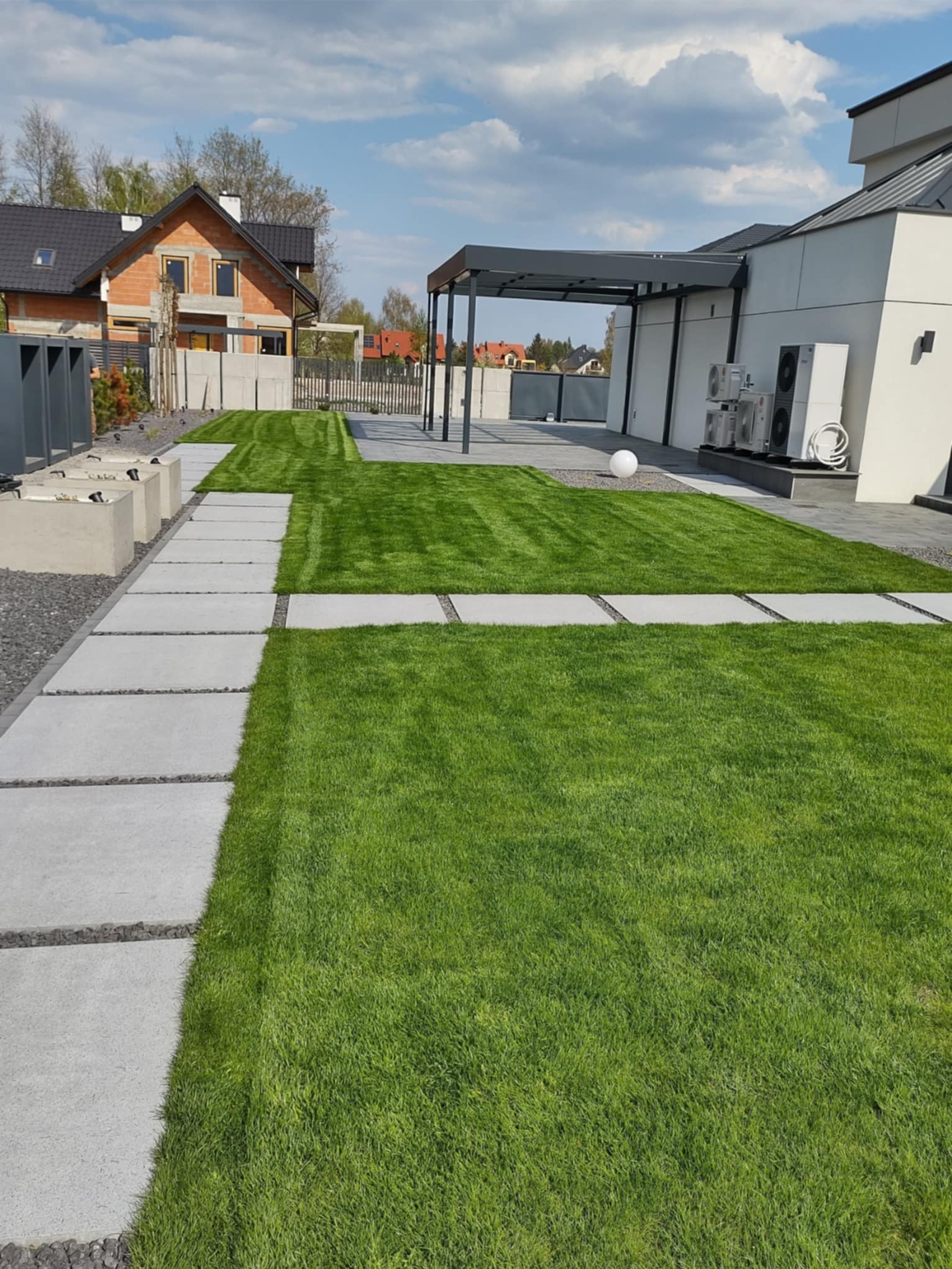 Nowoczesny ogród z równo przystrzyżonym trawnikiem, betonowymi płytami chodnikowymi i minimalistyczną pergolą. Widoczne elementy architektury domu i starannie dobrane rośliny.