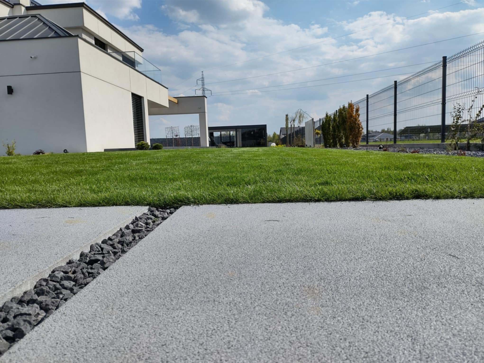 Nowoczesny ogród z równo przystrzyżonym trawnikiem, szarą ścieżką z kamieniami i minimalistyczną architekturą domu w tle, w słoneczny dzień.