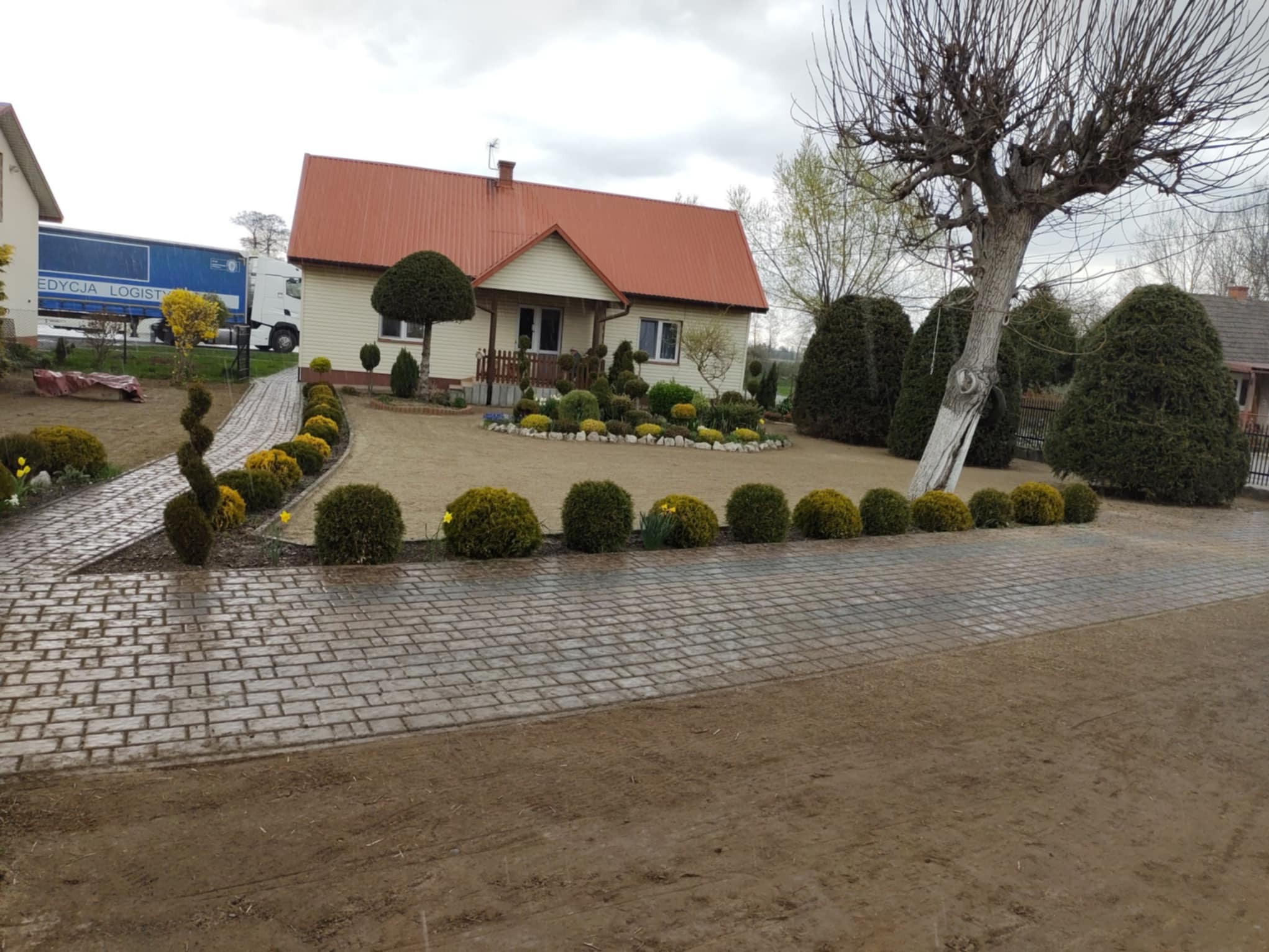 Uporządkowany ogród z równo przyciętymi krzewami i ścieżką z kostki brukowej prowadzącą do domu z czerwonym dachem. W tle widoczny samochód ciężarowy.