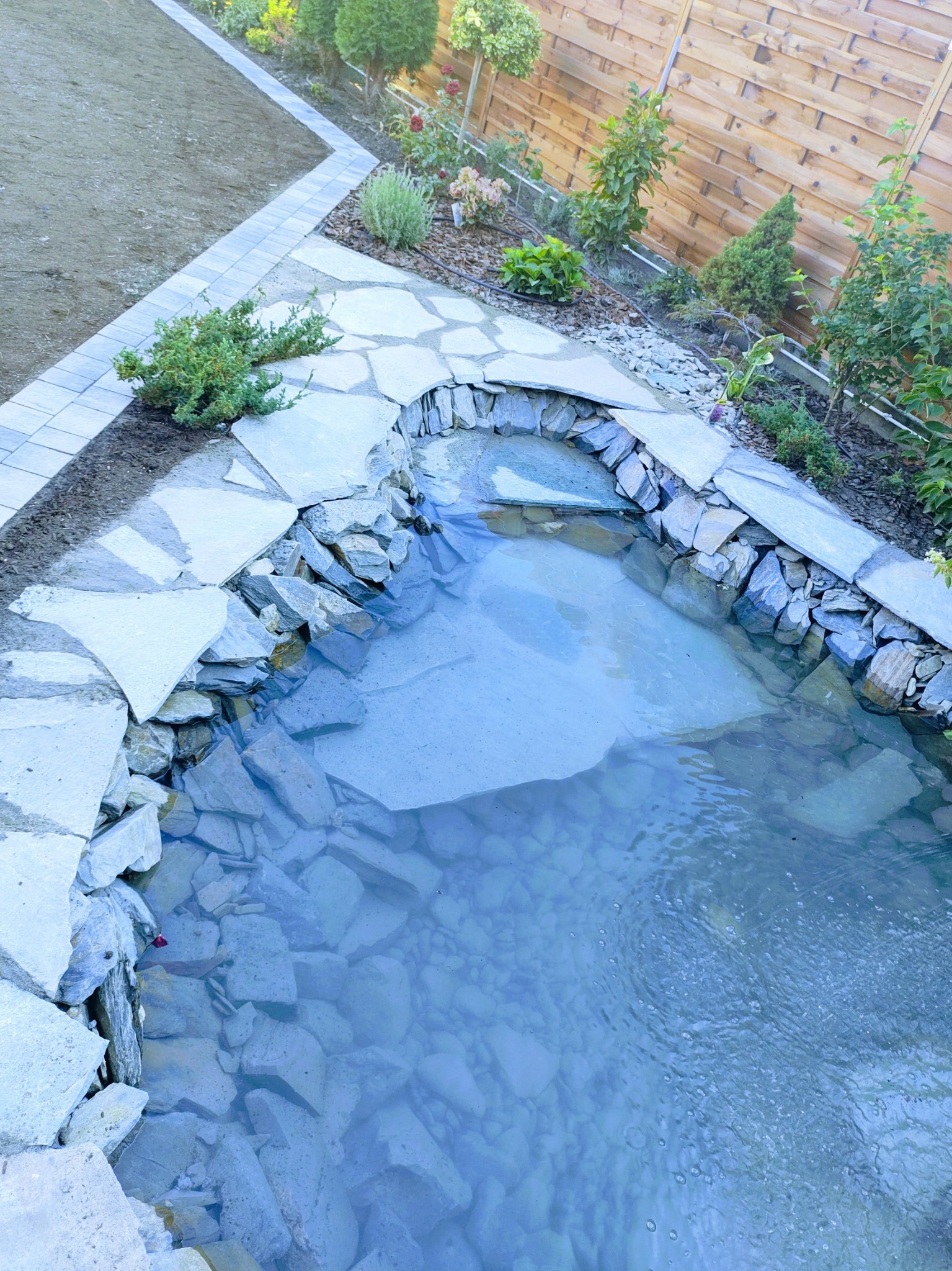 Oczko wodne z kamienną obudową i ścieżką z płyt w ogrodzie. W tle drewniany płot i świeżo założony trawnik. Widok z góry.