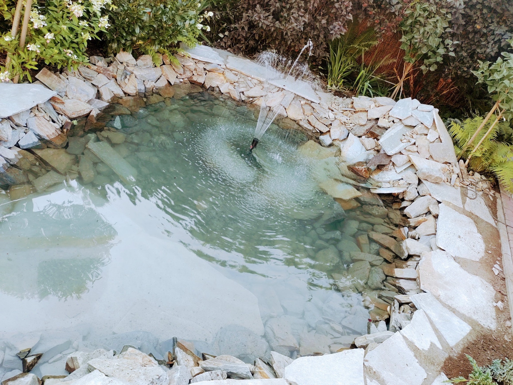 Oczko wodne z kamienną obudową i fontanną, otoczone zielenią. Widok z góry ukazuje przejrzystą wodę i kamienne dno. Element dekoracyjny ogrodu.