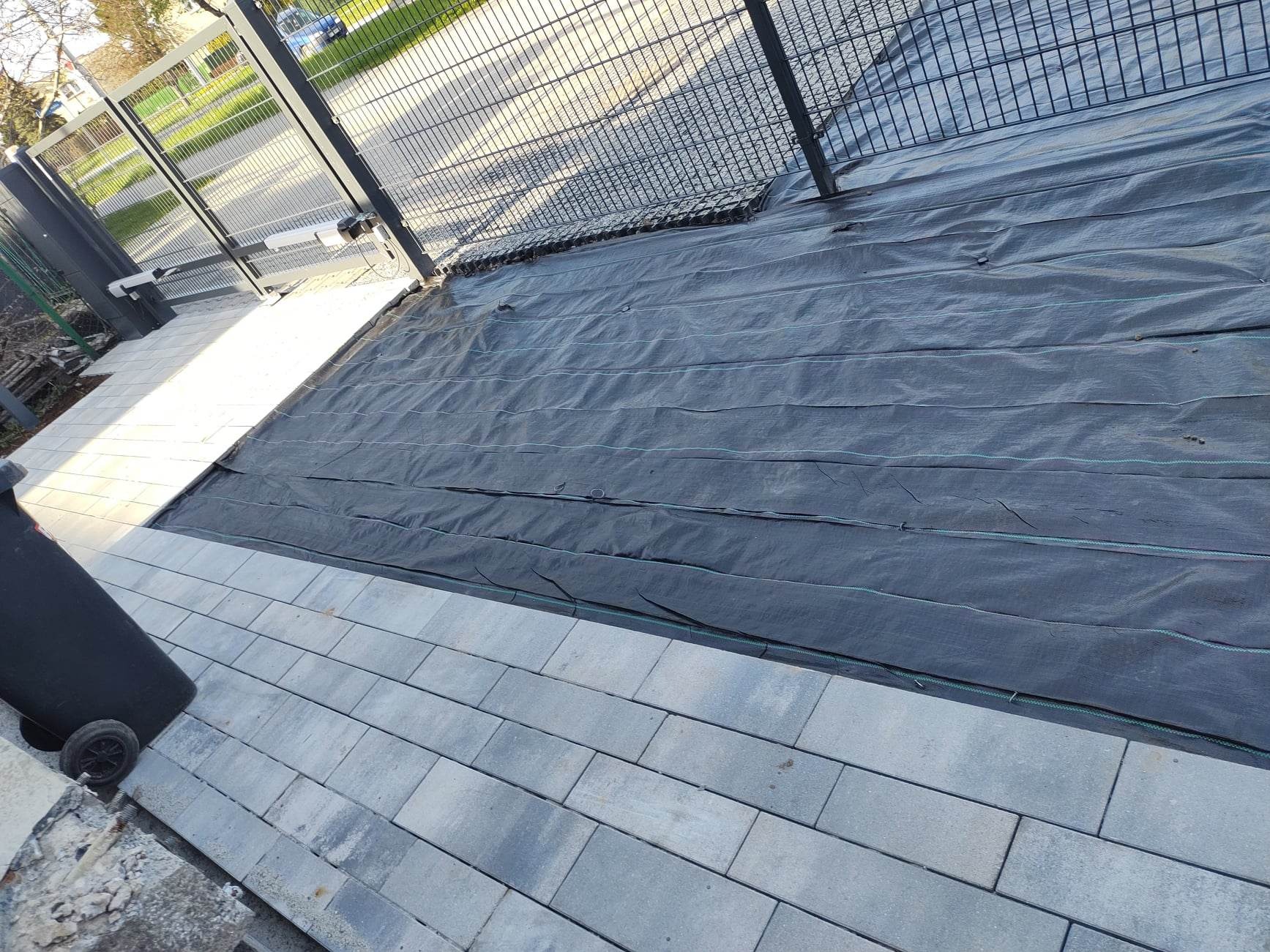 Układanie kostki brukowej przy ogrodzeniu, z widoczną czarną agrowłókniną. Częściowo ułożona nawierzchnia z szarych prostokątnych kostek.