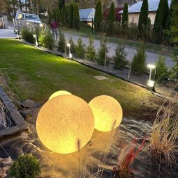 Unlimited Volt - Instalacje elektryczne - Wieczorny ogród z dwoma dużymi, teksturowanymi kulami świetlnymi w otoczeniu traw ozdobnych oraz rzędem minimalistycznych lamp słupkowych wzdłuż trawnika.