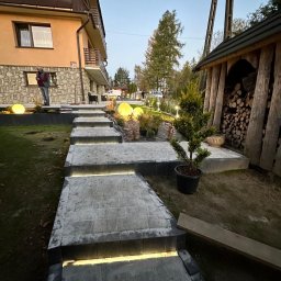 Unlimited Volt - Instalacje elektryczne - Schody zewnętrzne z kostki brukowej z wbudowanym oświetleniem LED, prowadzące do budynku mieszkalnego. Obok dekoracyjne kule świetlne i drewutnia o zmierzchu.
