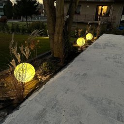 Unlimited Volt - Instalacje elektryczne - Trzy świecące, żółte kule ogrodowe oświetlające rabatę z trawami ozdobnymi, ułożone wzdłuż ścieżki pokrytej piaskiem, na tle domu o zmierzchu.