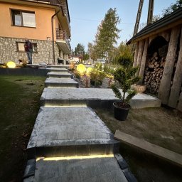 Unlimited Volt - Instalacje elektryczne - Podświetlane kamienne schody ogrodowe z wbudowanymi listwami LED, prowadzące do domu, otoczone kulistymi lampami i drewutnią, o zmierzchu.
