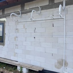 TUBERO - Białe rury instalacji zewnętrznej, częściowo owinięte folią ochronną, zamontowane na ścianie z białych bloczków. Widoczne ślady po montażu i szkic ołówkiem na ścianie.