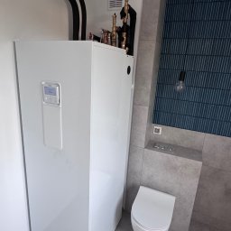 TUBERO - Biała pompa ciepła w nowoczesnej łazience z niebieską mozaiką na ścianie i podwieszaną toaletą. Widoczne rury instalacji i minimalistyczna lampa.