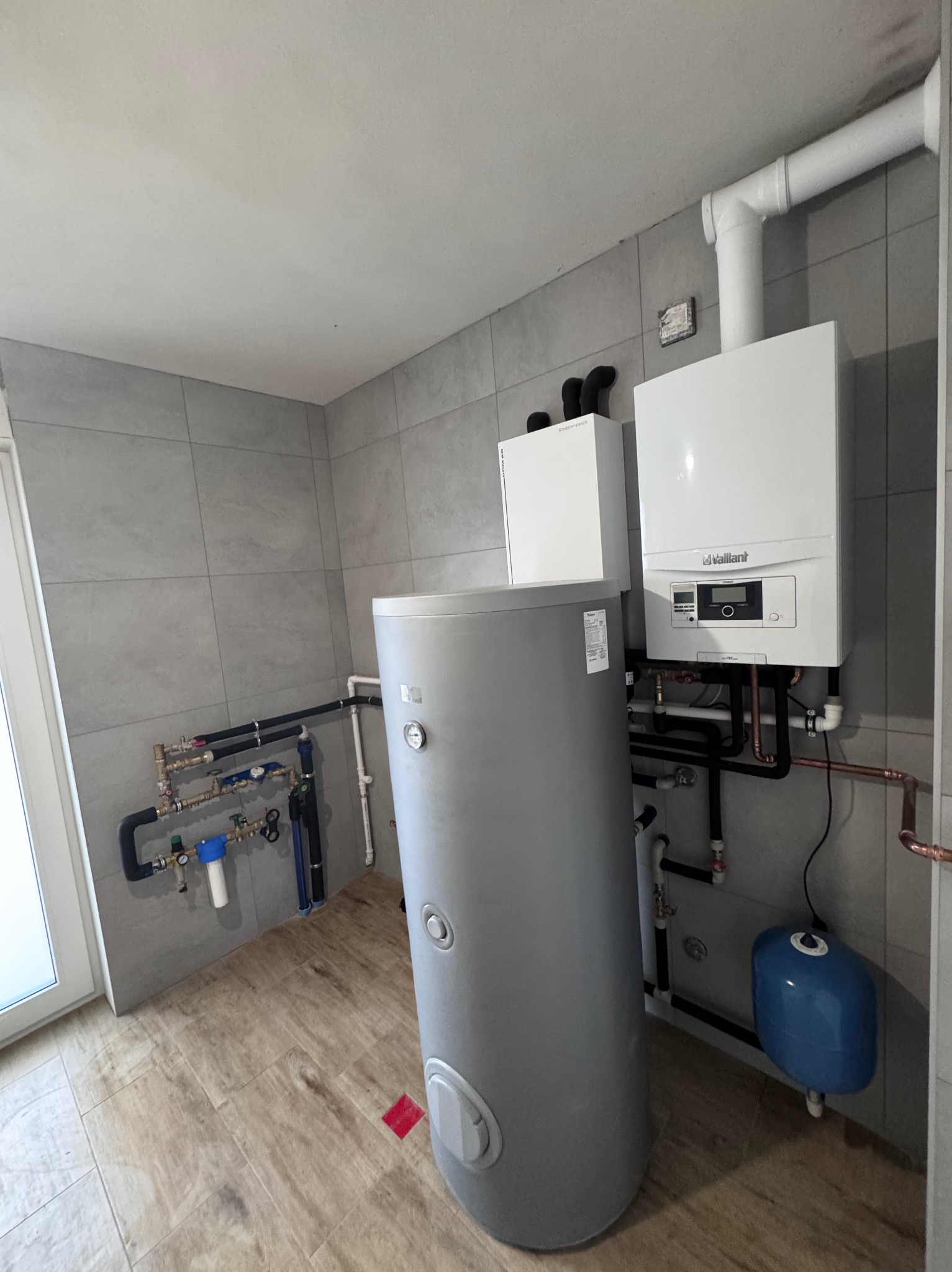 Nowoczesna kotłownia z szarym zasobnikiem ciepłej wody, białym piecem gazowym Vaillant i instalacją hydrauliczną na tle szarych płytek. Widoczny niebieski zbiornik wyrównawczy.