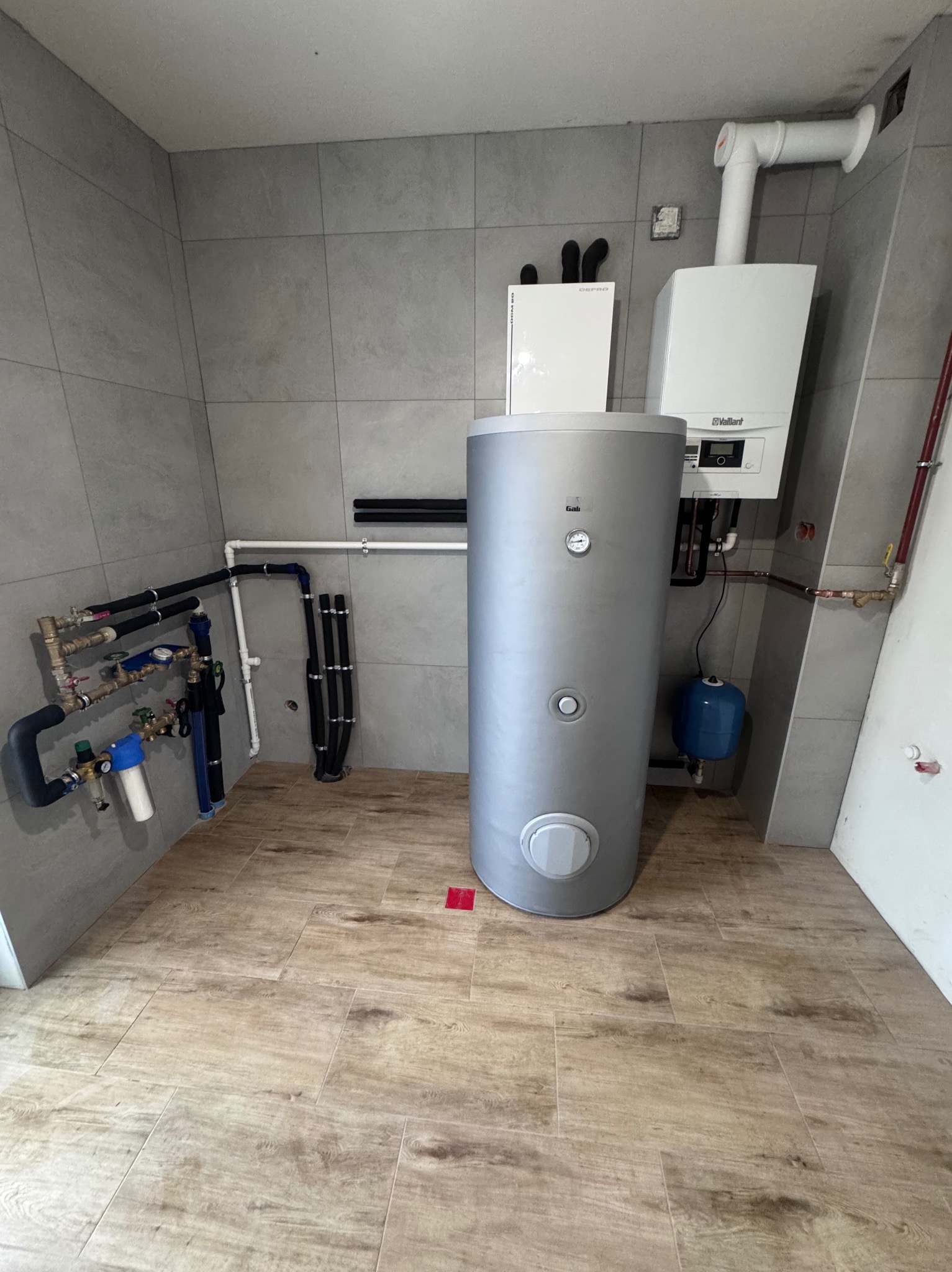 Szara kotłownia z piecem gazowym Vaillant, zasobnikiem ciepłej wody i instalacją hydrauliczną. Nowoczesne płytki na ścianach i podłodze.