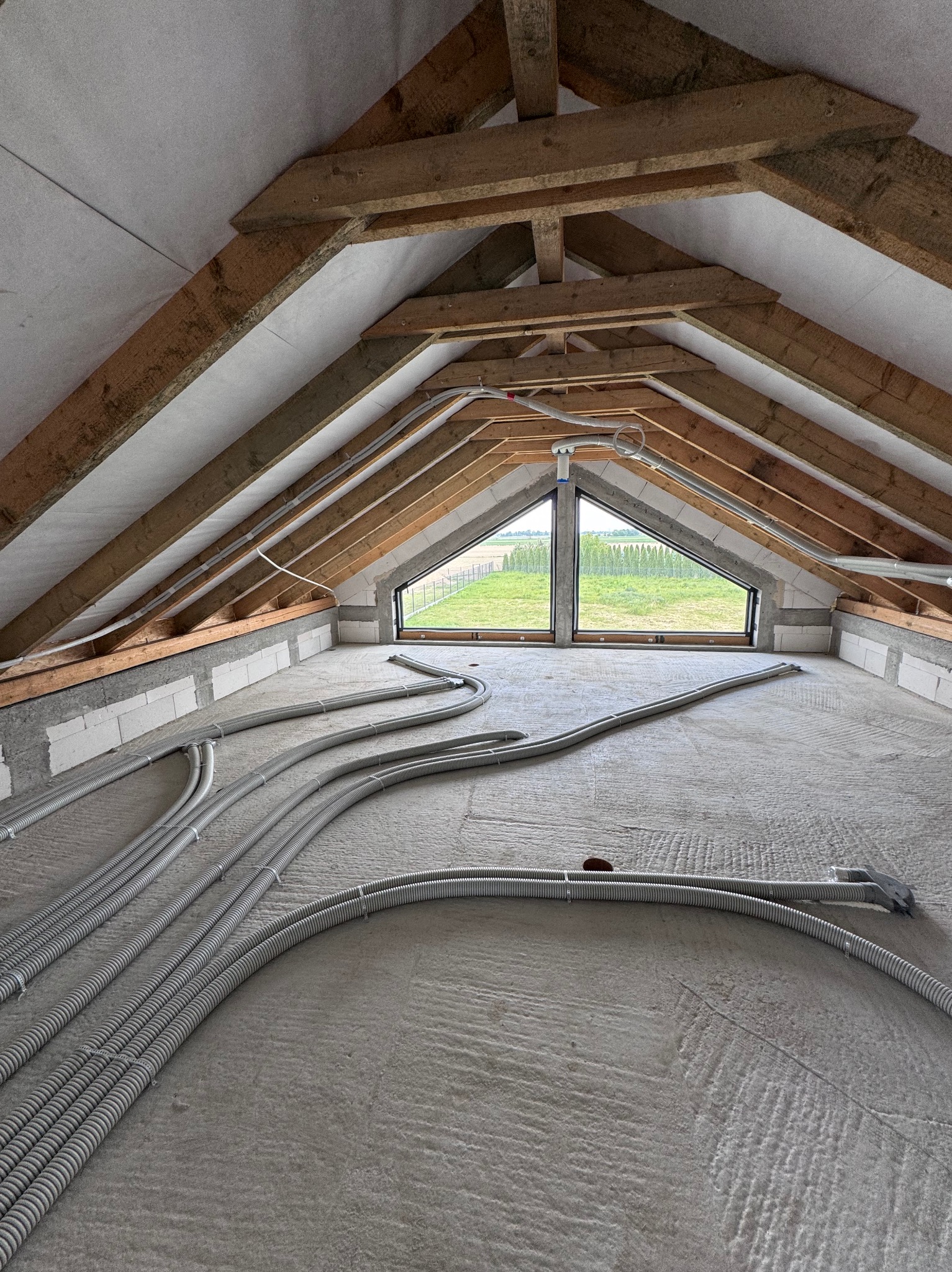 Poddasze z oknami i widoczną instalacją przewodów w peszlach, ułożonych na izolacji. Drewniana konstrukcja dachu i surowe ściany z betonu komórkowego.