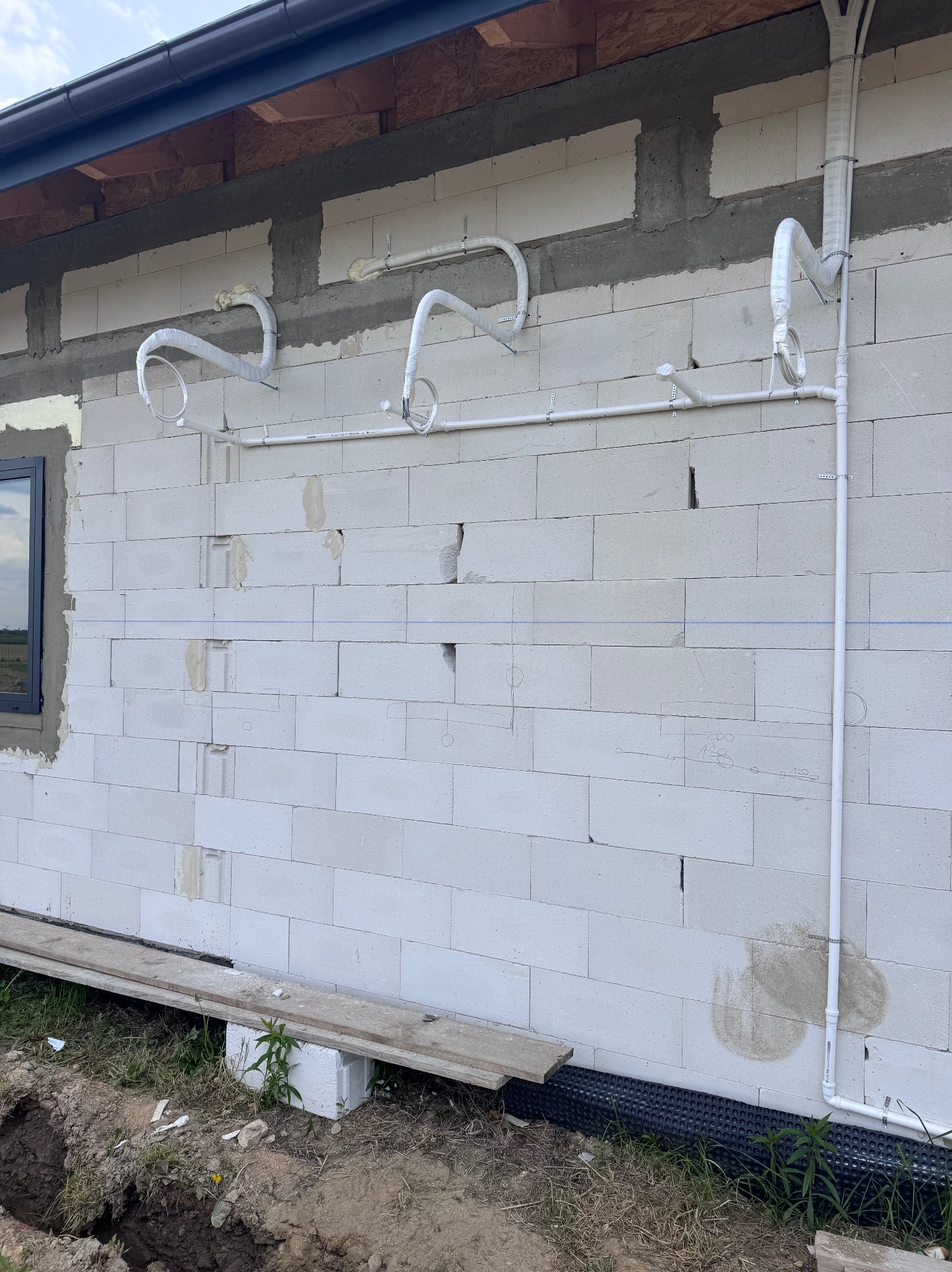 Białe rury instalacji zewnętrznej, częściowo owinięte folią ochronną, zamontowane na ścianie z białych bloczków. Widoczne ślady po montażu i szkic ołówkiem na ścianie.