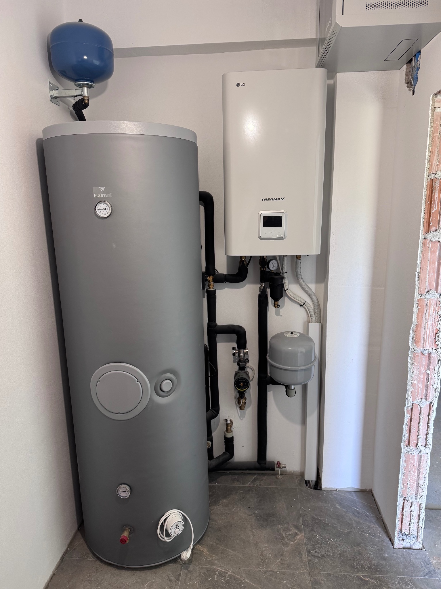 Szary, cylindryczny zasobnik ciepłej wody Calmet z niebieskim naczyniem wzbiorczym i biały piec LG Therma V, zamontowane na ścianie w pomieszczeniu z szarą podłogą.