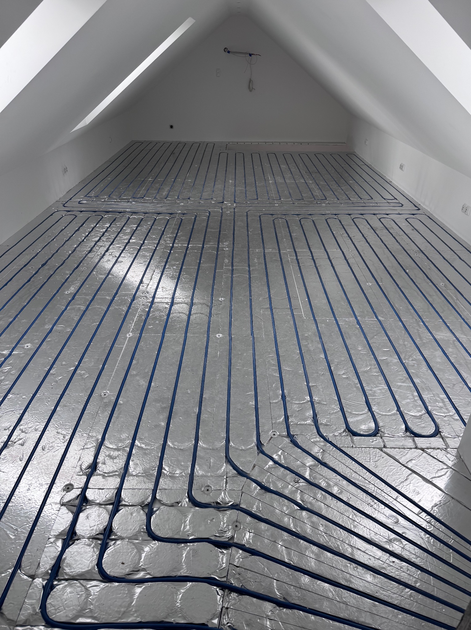 Instalacja ogrzewania podłogowego w poddaszu: niebieskie rurki na srebrnej folii izolacyjnej, widok z góry, surowe wykończenie, skosy dachu.