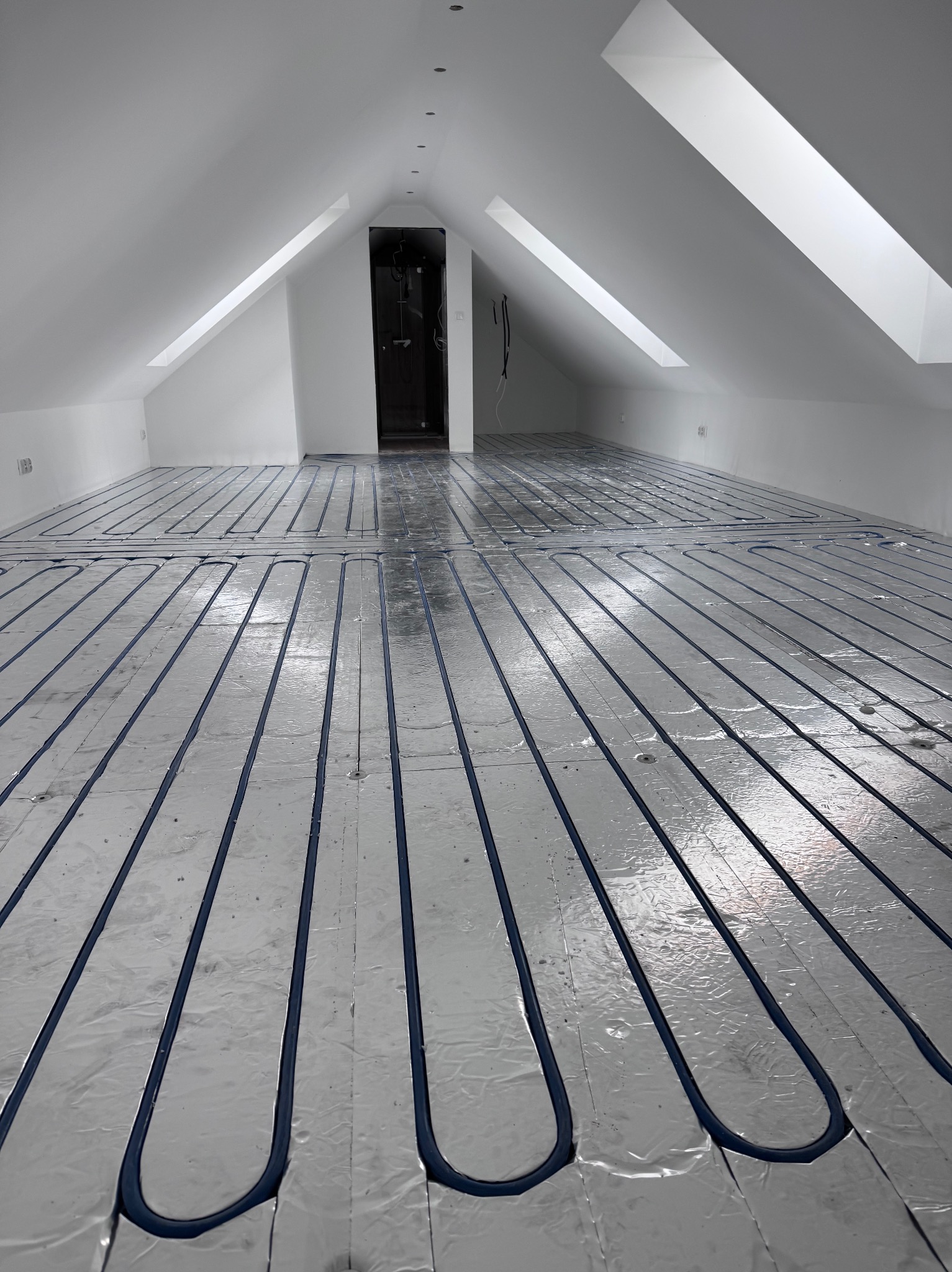 Ułożone rury ogrzewania podłogowego na srebrnej folii w jasnym, przestronnym poddaszu z oknami dachowymi i widocznym wejściem do pomieszczenia.