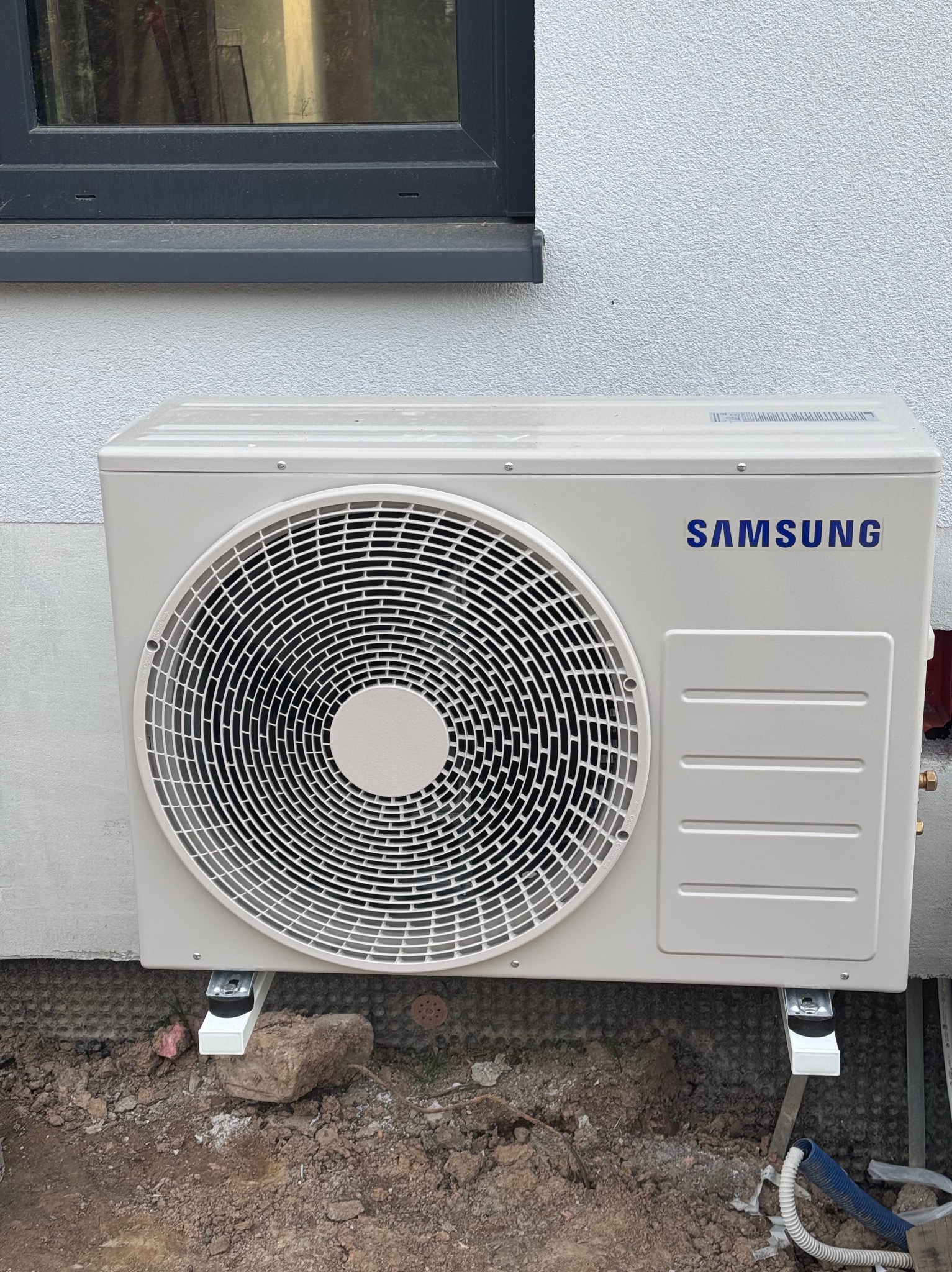 Zamontowana jednostka zewnętrzna klimatyzacji Samsung na elewacji budynku, widoczne przewody instalacyjne i fragment okna. Ujęcie z poziomu gruntu.