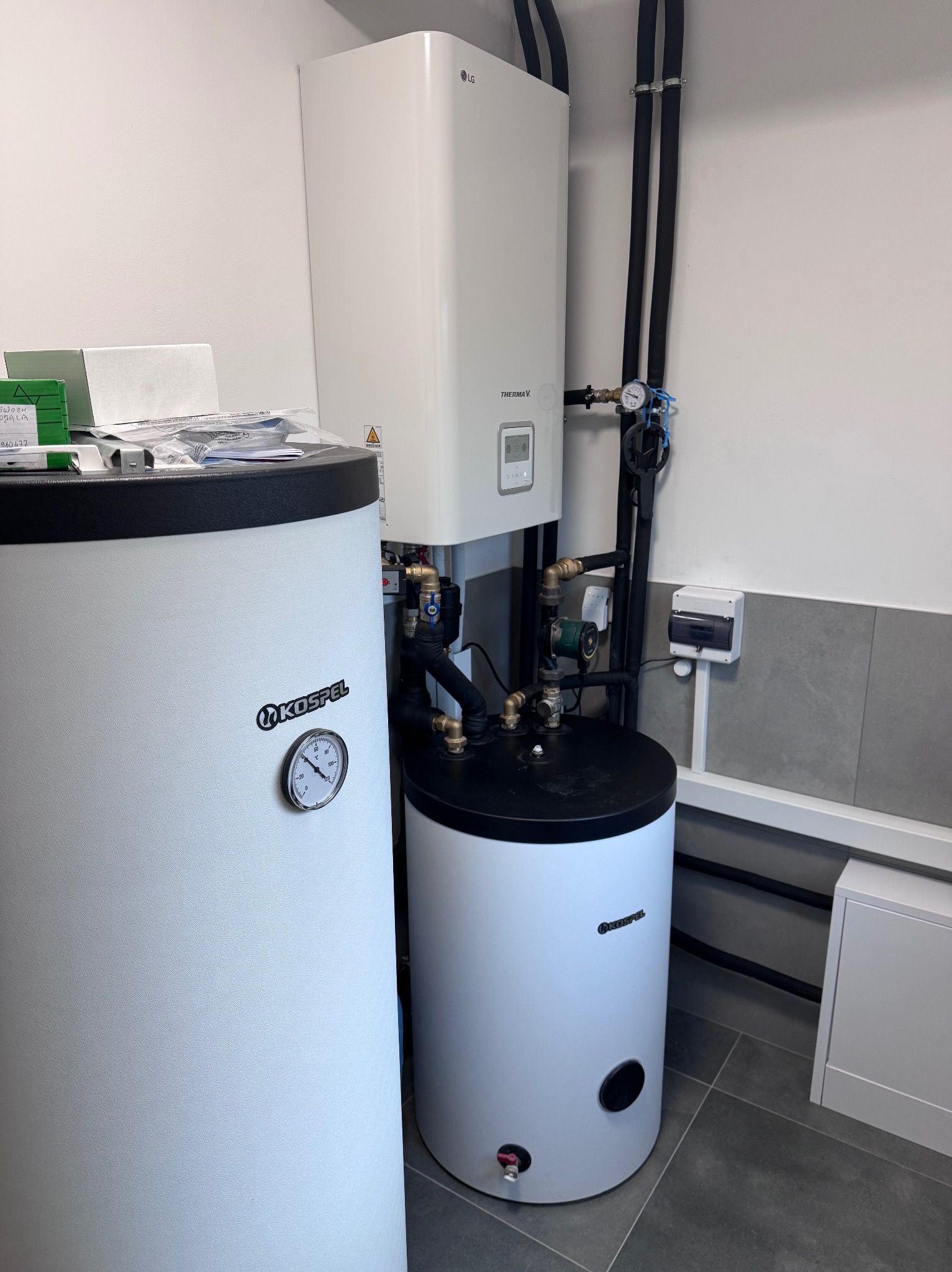 Biały piec CO marki LG Thermav i zbiornik Kospel z manometrem, połączone rurami w schludnej kotłowni z szarą podłogą i białymi ścianami.