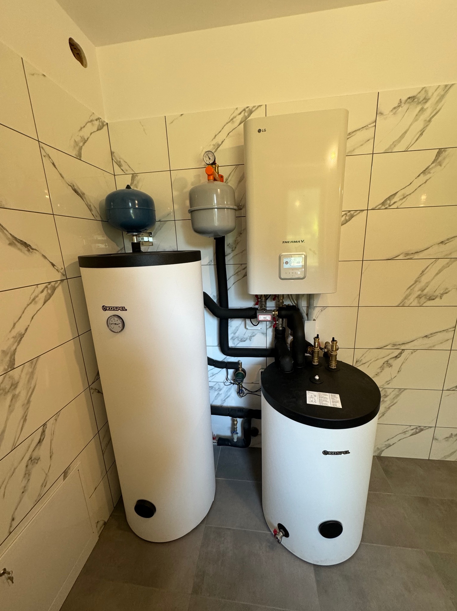 Biały piec gazowy LG i dwa białe zbiorniki Kospel z czarnymi elementami, połączone rurami w pomieszczeniu z marmurowymi płytkami i szarą podłogą.