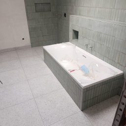 Nowoczesna łazienka w trakcie wykańczania: biała wanna obudowana szarą mozaiką, ściany wyłożone pionowymi, szarymi płytkami. Podłoga z jasnych, nakrapianych płytek.