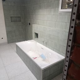 Nowoczesna łazienka w trakcie wykończenia: wanna obudowana płytkami, ściany wyłożone podłużnymi, zielonkawymi płytkami. Widoczne przygotowane otwory na armaturę.