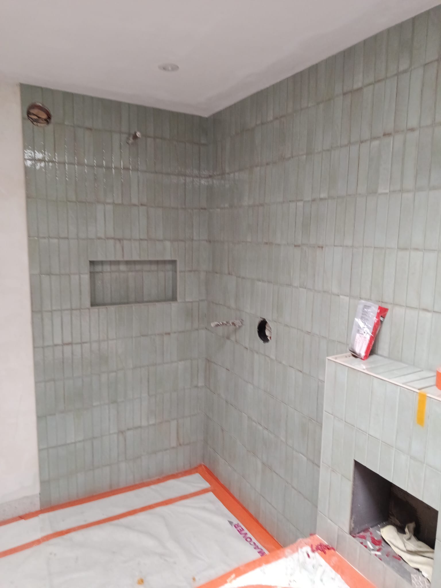 Ściany w łazience wyłożone pionowymi, zielonkawymi płytkami. Widoczna wnęka w ścianie i otwór na instalację. Podłoga zabezpieczona folią ochronną.