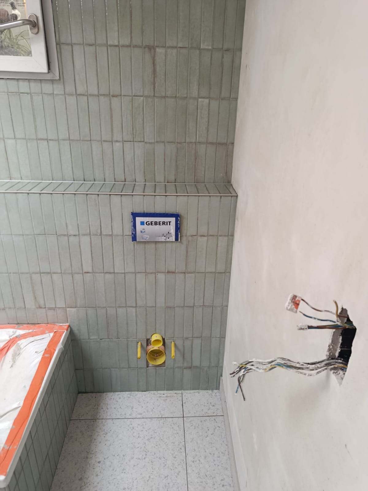 Ściana łazienki z ułożonymi, wąskimi, zielonkawymi płytkami. Widoczna instalacja Geberit i otwór w ścianie z wystającymi kablami elektrycznymi.