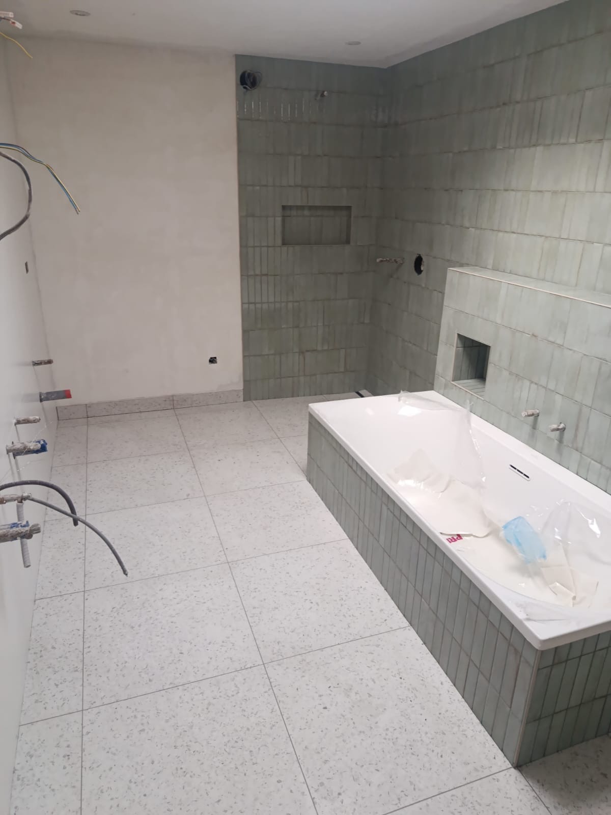 Wnętrze łazienki w trakcie wykańczania. Widoczna wanna obudowana płytkami, ściany częściowo wyłożone płytkami w odcieniach szarości, instalacje wodne i elektryczne.