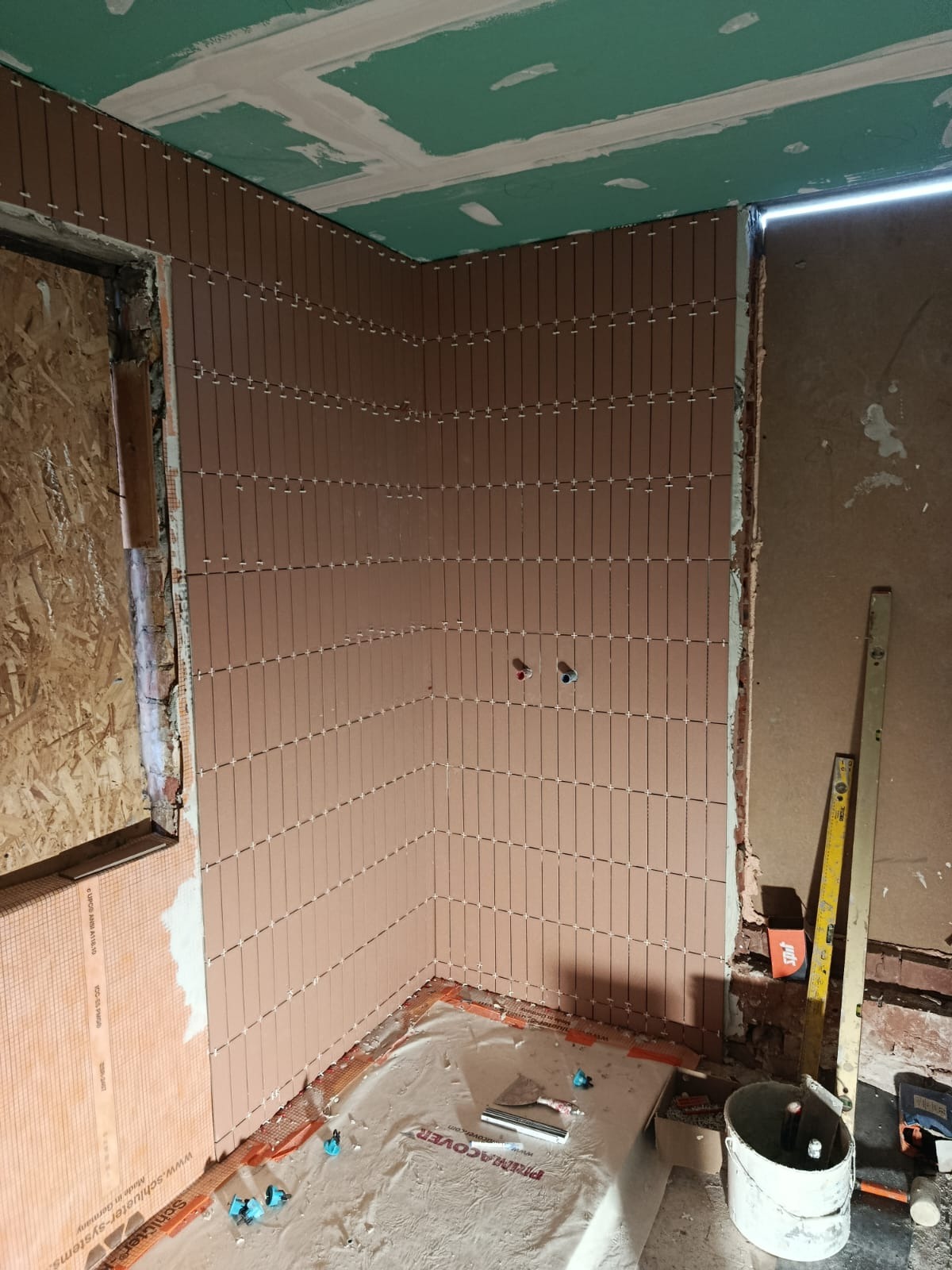 Układanie wąskich, brązowych płytek na ścianie łazienki, widoczne krzyżyki dystansowe, fragment instalacji wodnej, poziomica i wiadro. Prace glazurnicze w toku.