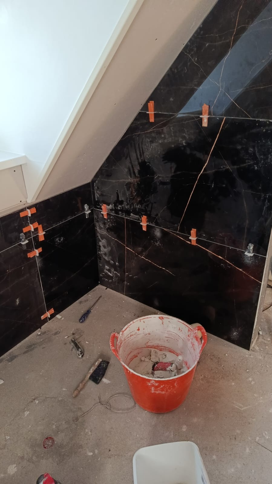 Układanie czarnych, marmurkowych płytek w narożniku pomieszczenia. Widoczne pomarańczowe krzyżyki dystansowe i narzędzia glazurnicze, w tym wiadro z zaprawą.