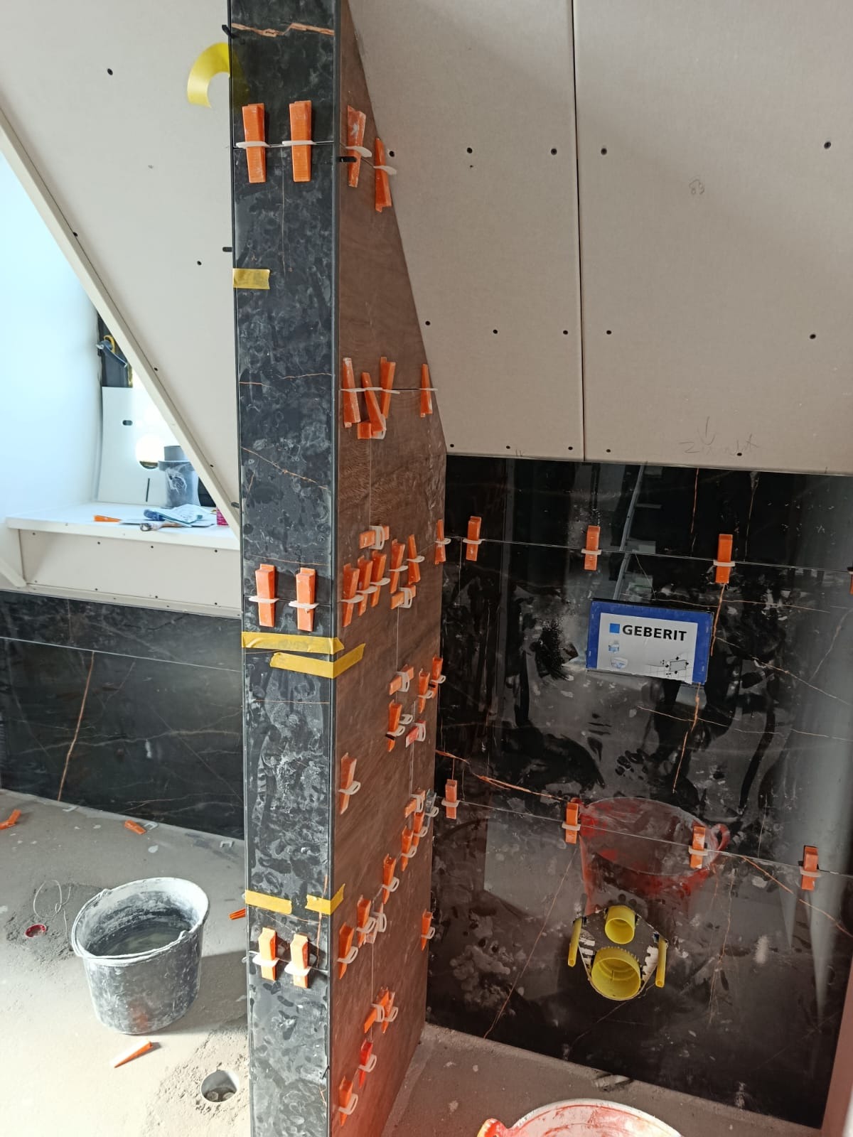 Montaż płytek imitujących marmur w łazience z użyciem klipsów poziomujących. Widoczna instalacja Geberit i fragment sufitu ze skosem. Prace w toku.