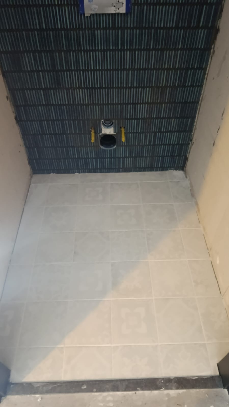 Łazienka w trakcie remontu: ułożone płytki podłogowe w jasnym odcieniu i dekoracyjne, ciemne płytki na ścianie, widoczne elementy instalacji sanitarnej.