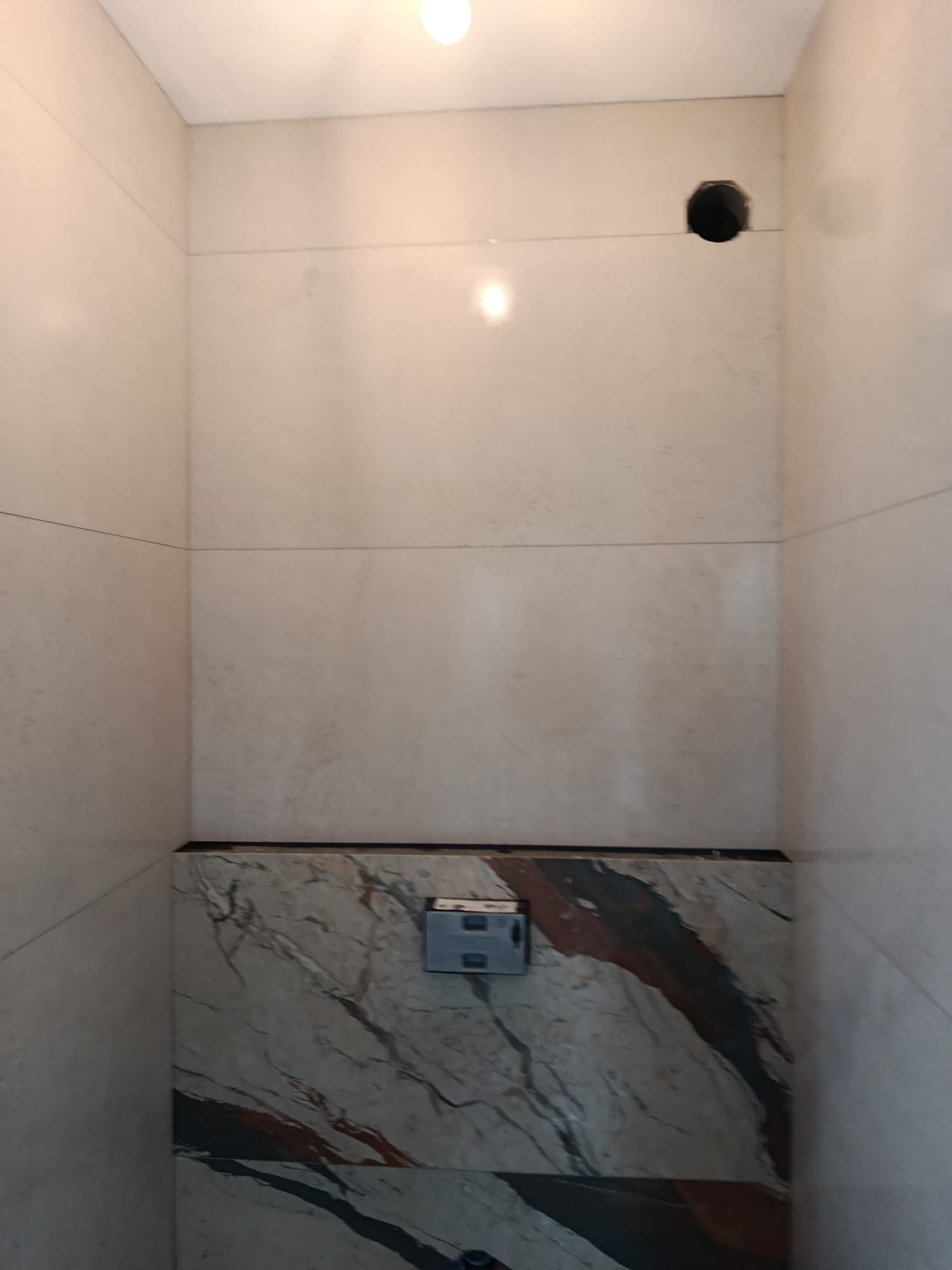 Wykończona ściana łazienki z beżowymi płytkami i marmurowym akcentem. Widoczny otwór na instalację. Minimalistyczny design, jasne oświetlenie.