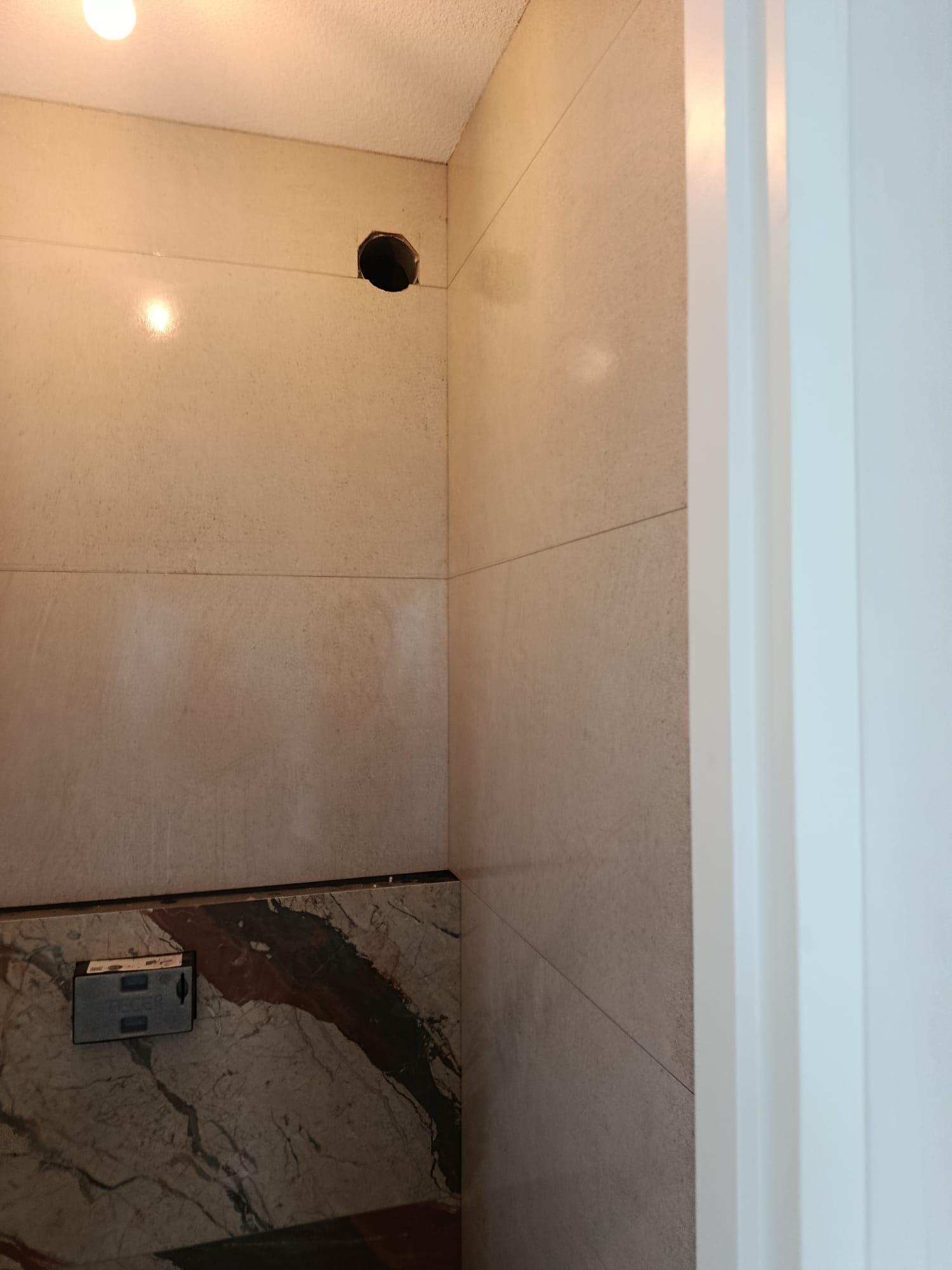 Fragment łazienki w trakcie wykańczania: beżowe płytki, marmurowy cokół, otwór w ścianie. Widoczna biała futryna drzwi. Oświetlenie sufitowe.