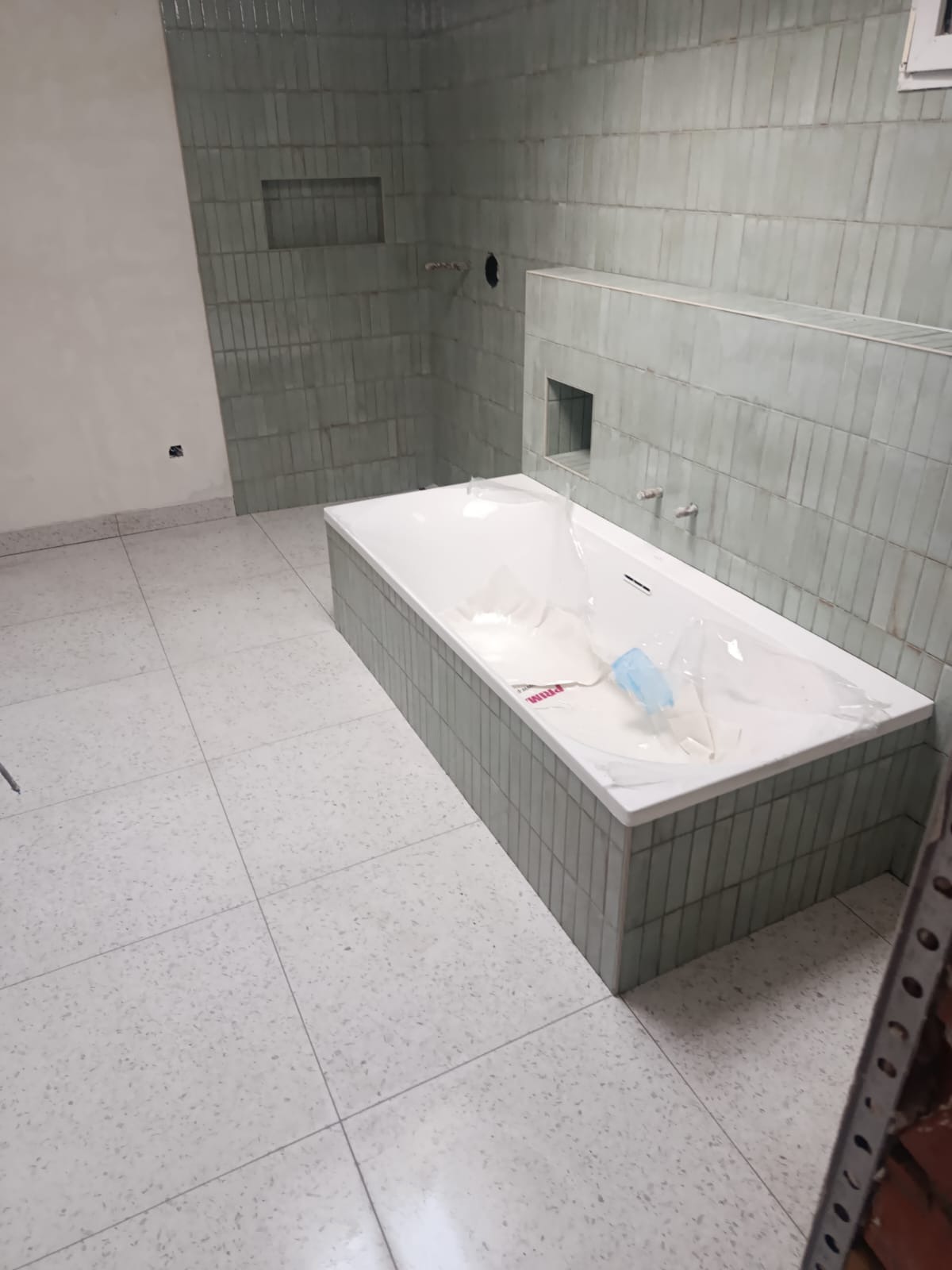 Nowoczesna łazienka w trakcie wykańczania: biała wanna obudowana szarą mozaiką, ściany wyłożone pionowymi, szarymi płytkami. Podłoga z jasnych, nakrapianych płytek.