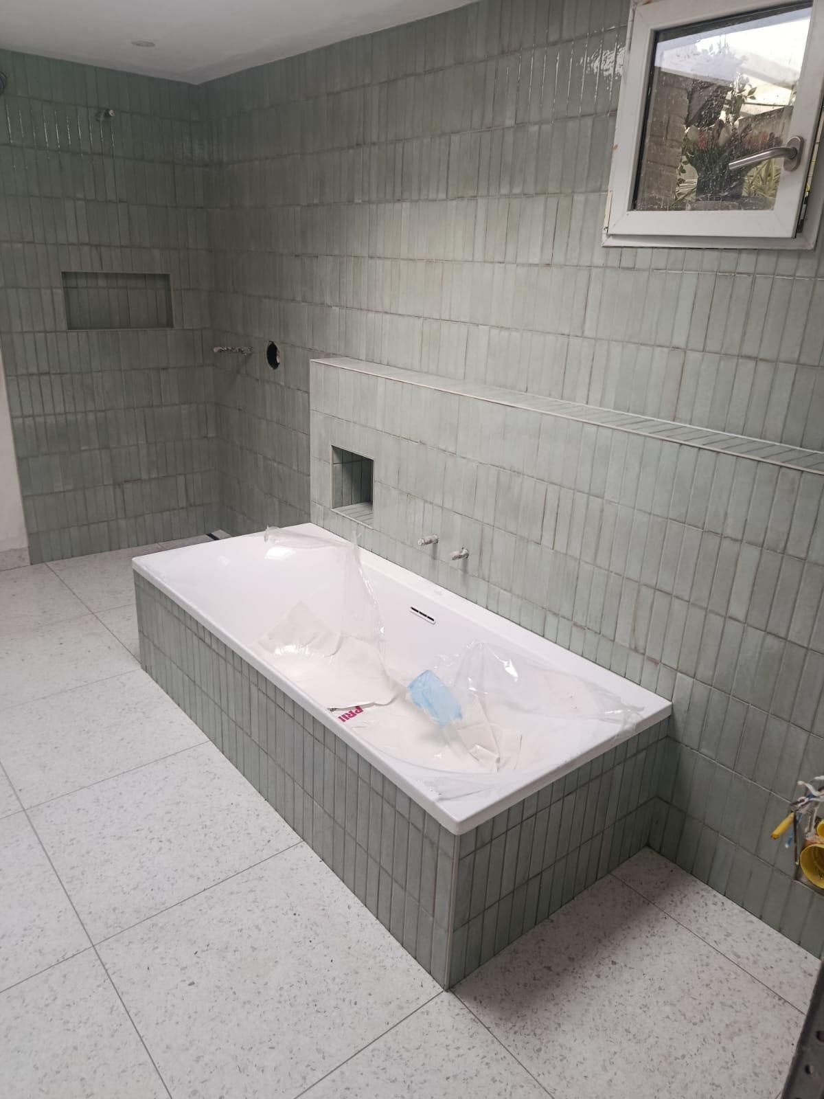 Wykończona łazienka z wanną obłożoną płytkami w odcieniach szarości. Na ścianach pionowe, wąskie płytki. Podłoga wyłożona jasnymi płytkami z delikatnym wzorem.