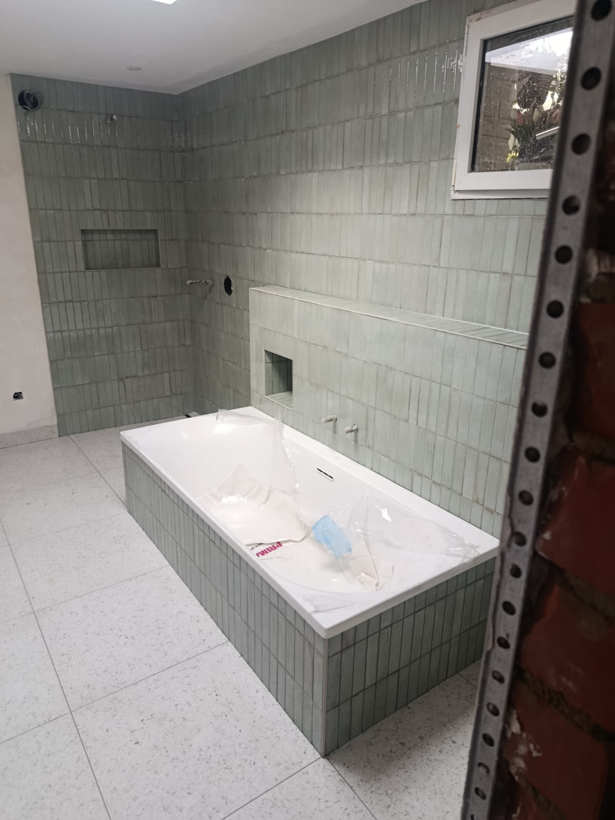 Nowoczesna łazienka w trakcie wykończenia: wanna obudowana płytkami, ściany wyłożone podłużnymi, zielonkawymi płytkami. Widoczne przygotowane otwory na armaturę.