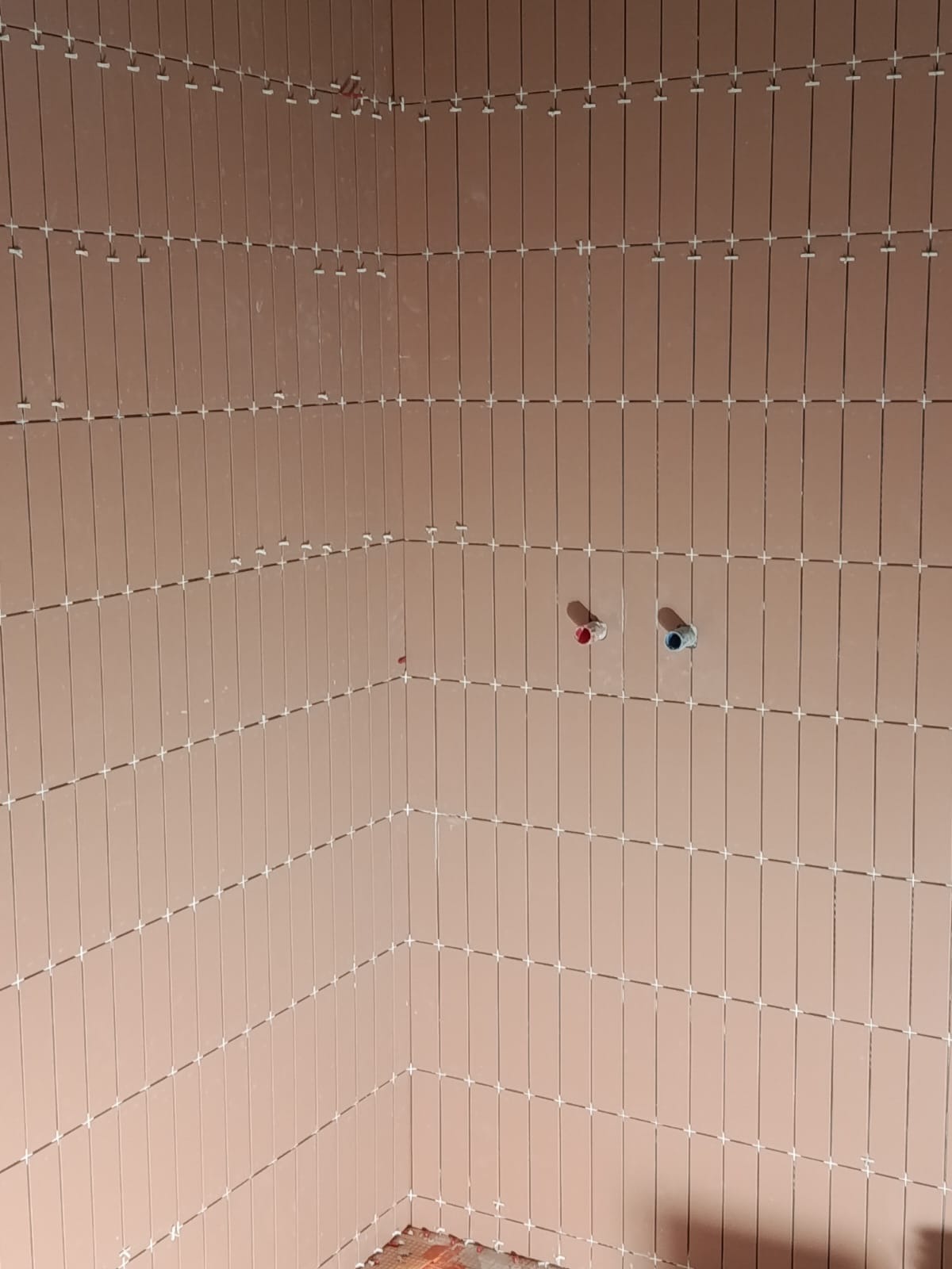 Łazienka w trakcie wykańczania: pionowo ułożone, podłużne, różowe płytki z białymi krzyżykami dystansowymi. Widoczne wyprowadzenia instalacji wodnej.