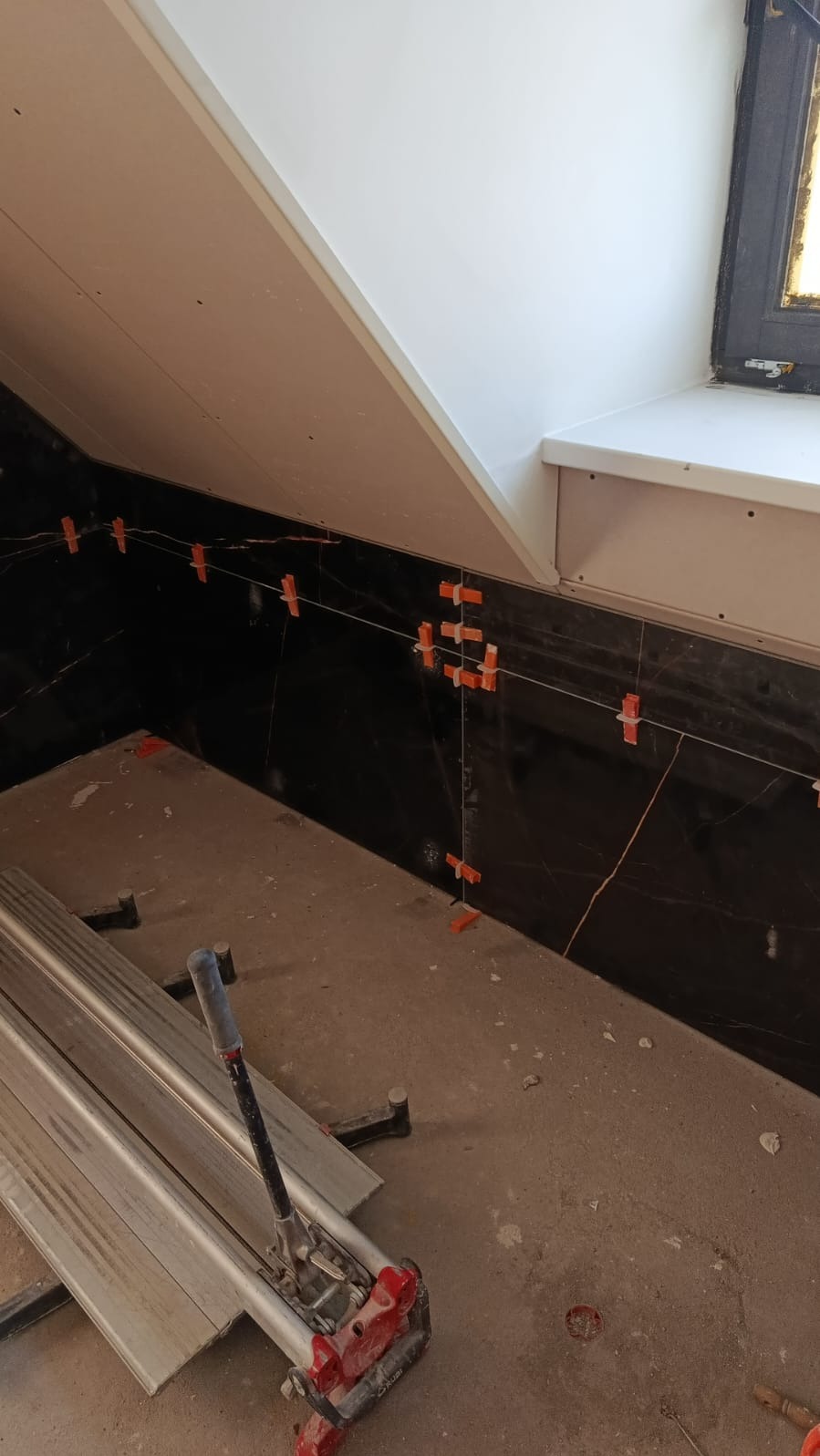 Częściowo wykończona ściana z czarnych płytek imitujących marmur, z pomarańczowymi krzyżykami dystansowymi i narzędziem do cięcia płytek na podłodze.