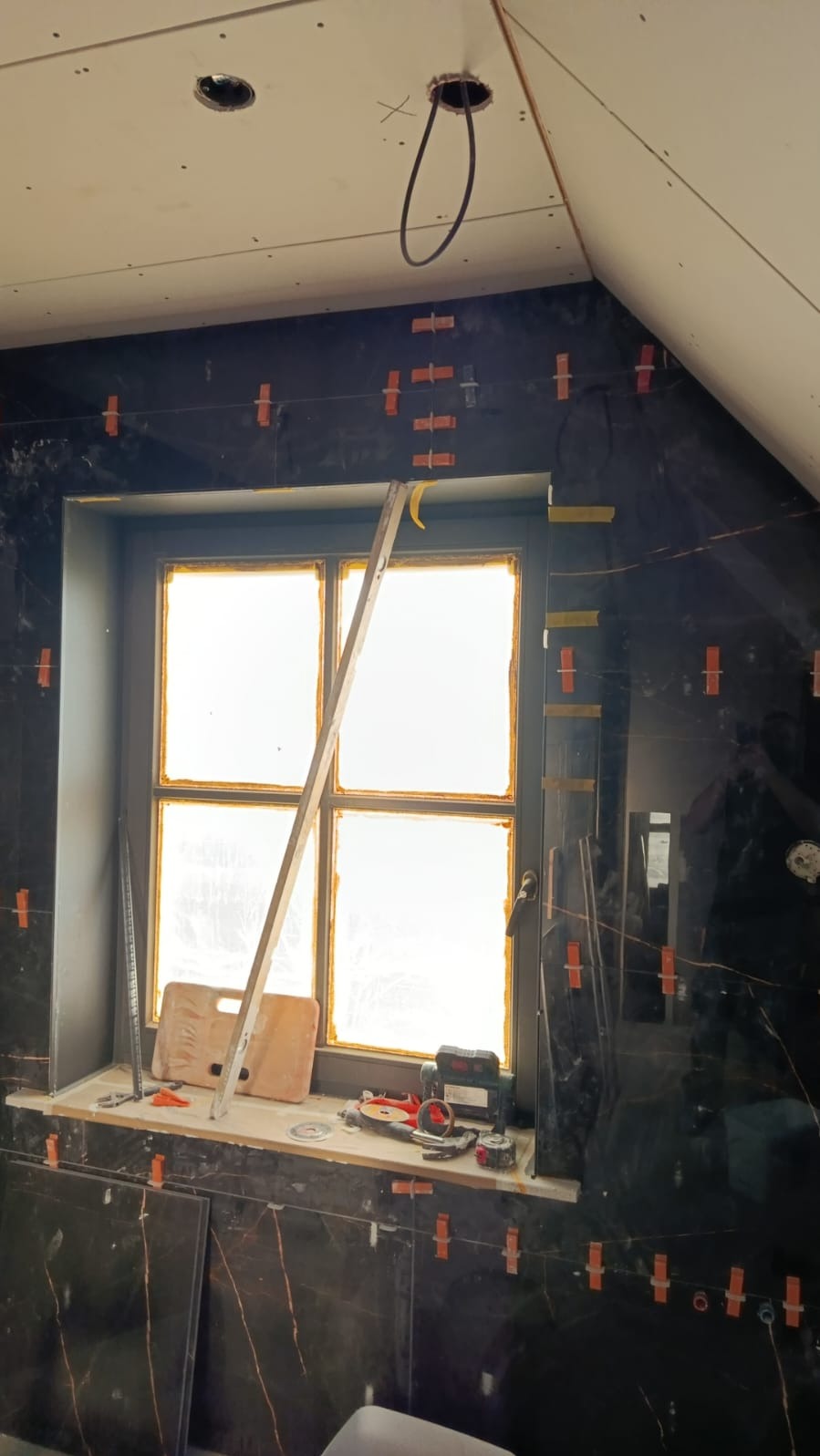 Ciemne, marmurowe płytki na ścianie w trakcie montażu, z użyciem krzyżyków dystansowych. Okno zabezpieczone folią, widoczne narzędzia glazurnicze i poziomica.