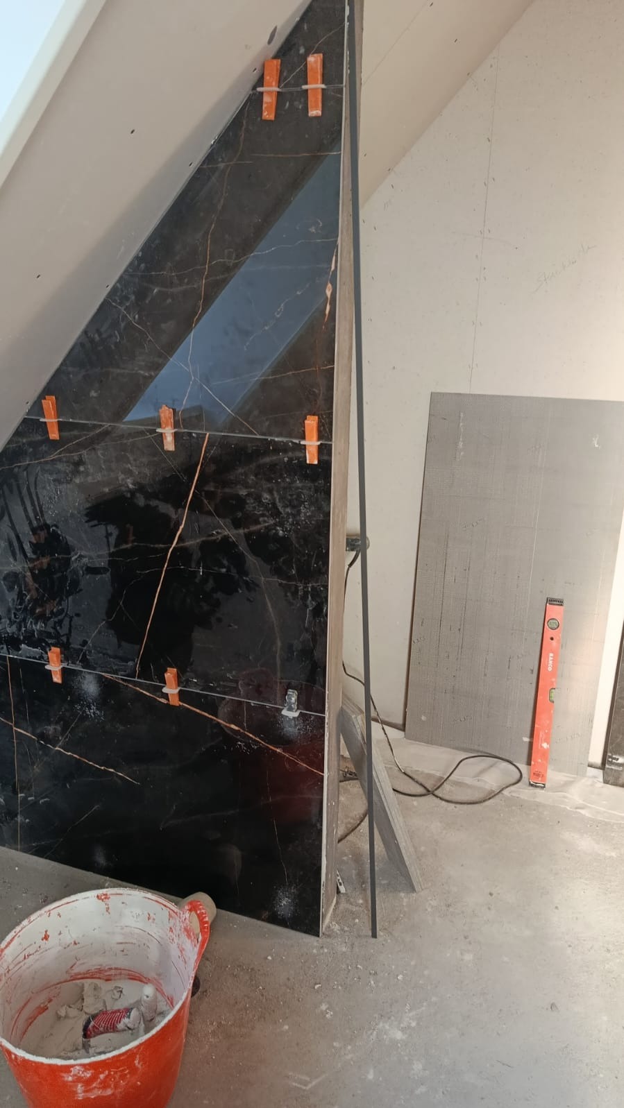 Ukończona ściana z czarnych, marmurkowych płytek w skosie poddasza, z pomarańczowymi krzyżykami dystansowymi. Wiadro z zaprawą i poziomica w tle.