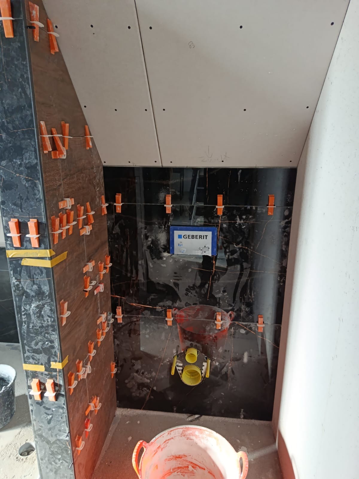 Montaż czarnych płytek z pomarańczowymi krzyżykami dystansowymi w łazience. Widoczna instalacja podtynkowa Geberit i płyta karton-gips na suficie. Ujęcie z dołu.