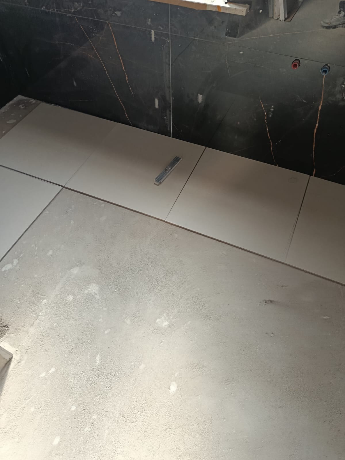 Układanie jasnych płytek podłogowych w łazience z ciemnymi, marmurkowymi płytkami ściennymi. Poziomica leży na płytkach.